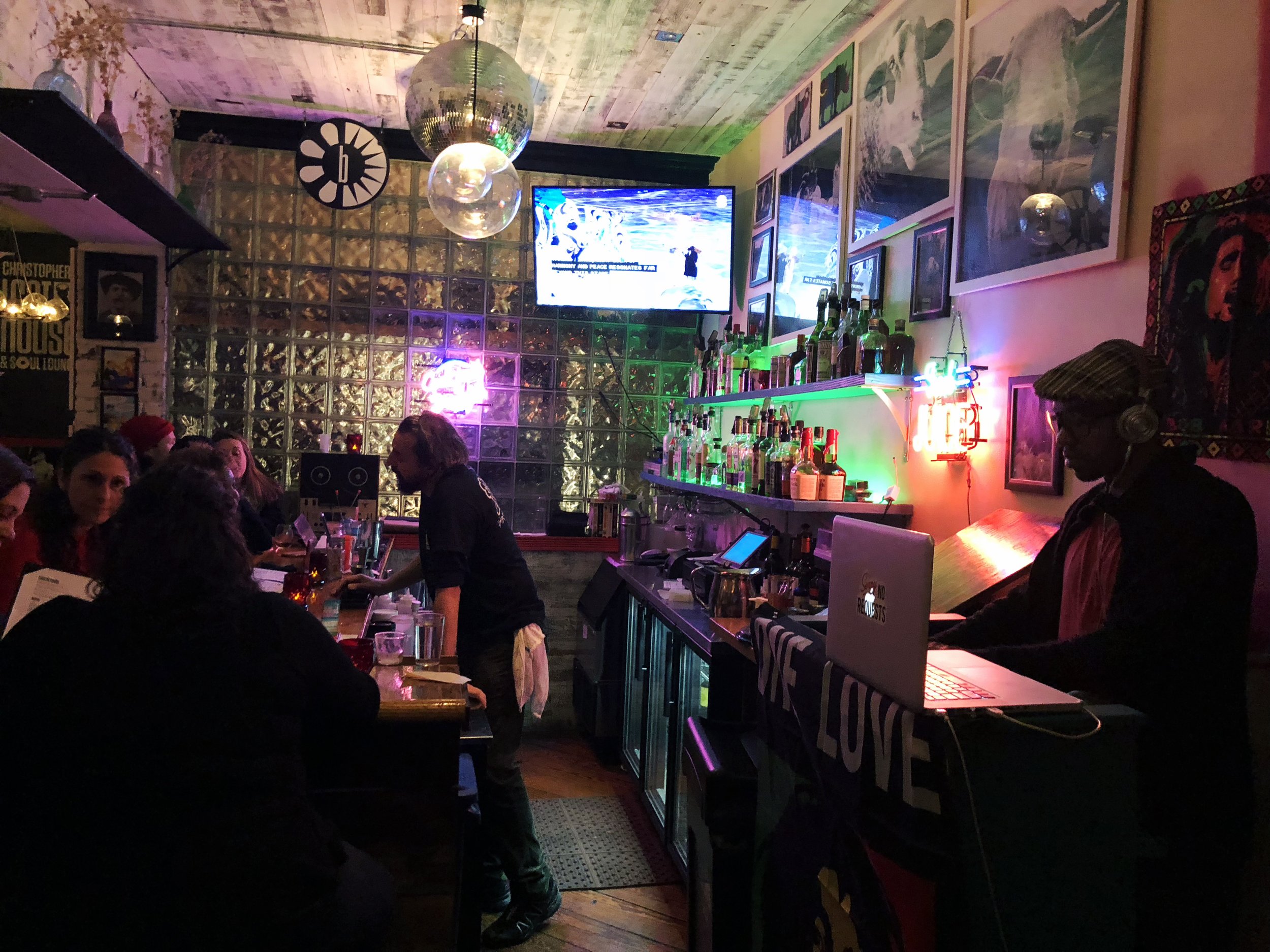 Crowded bar at Carmine St with a DJ, colorful lighting, artwork, and neighborhood guests in New York City.