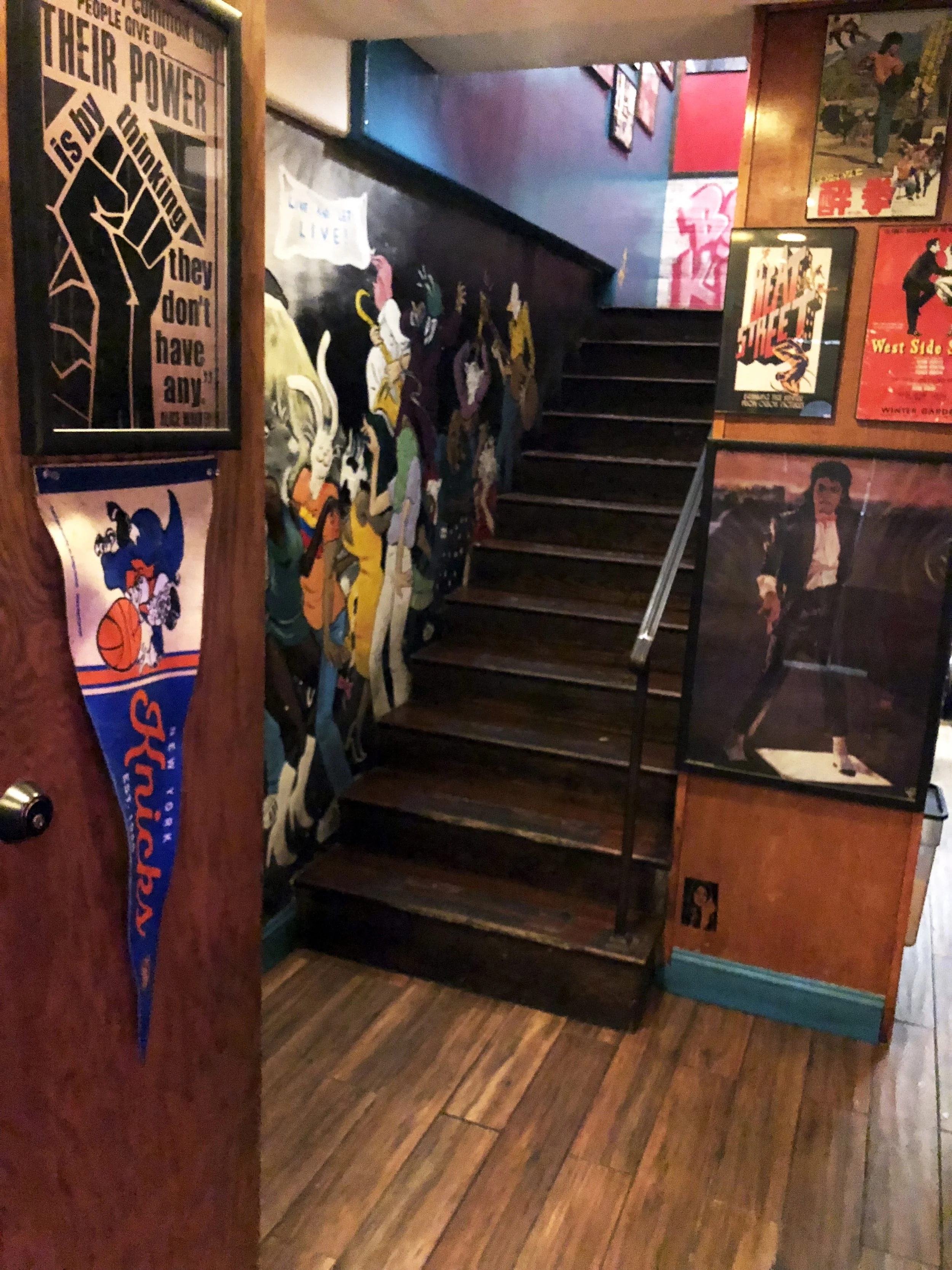 Stairway inside Carmine St lined with murals, posters, sports memorabilia, and New York cultural references.