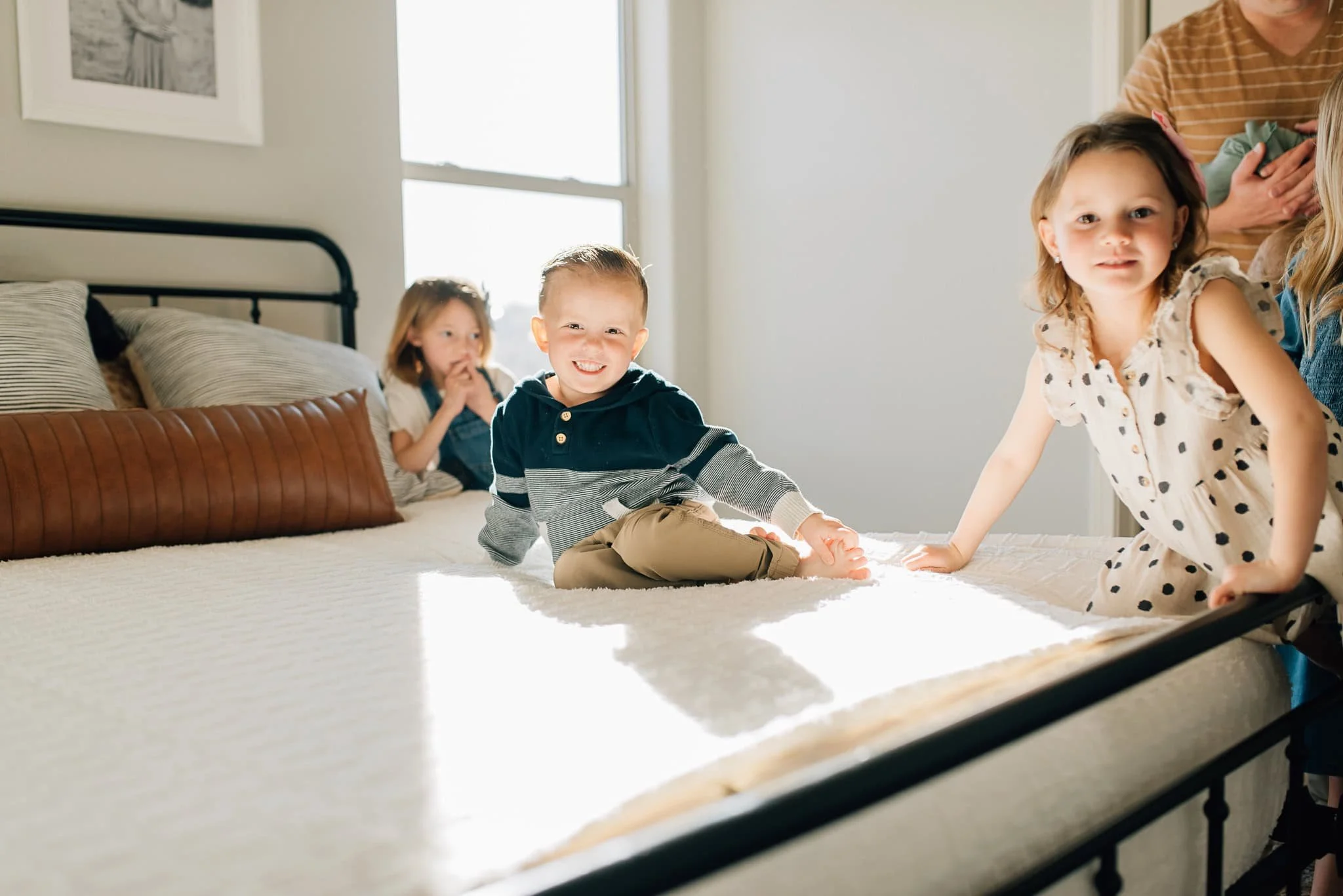little boy on bed smiles at camera while sisters climb on bed
