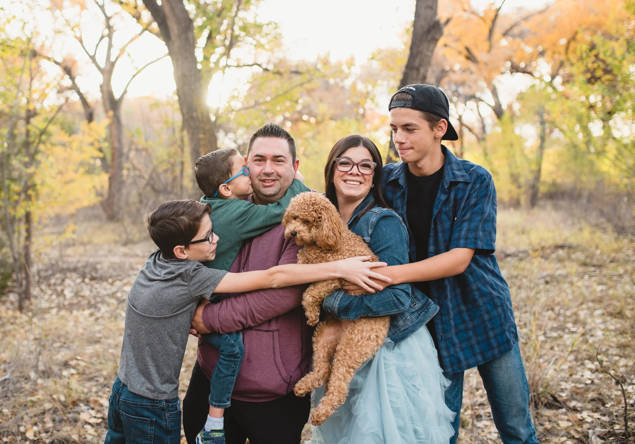 Family with three boys and dog hugging with fall leaves behind them