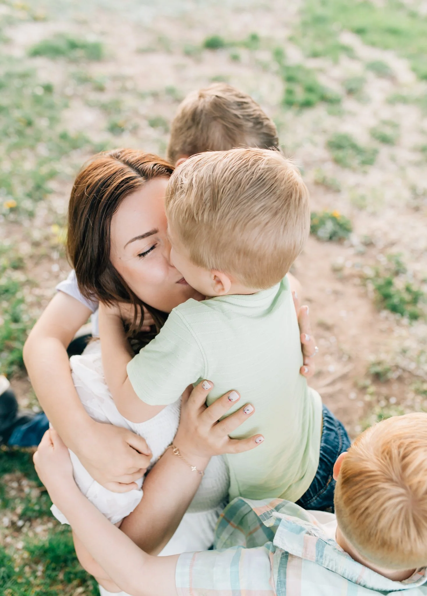 Mom sitting in grass with two kids in lap and one hugging her from behind