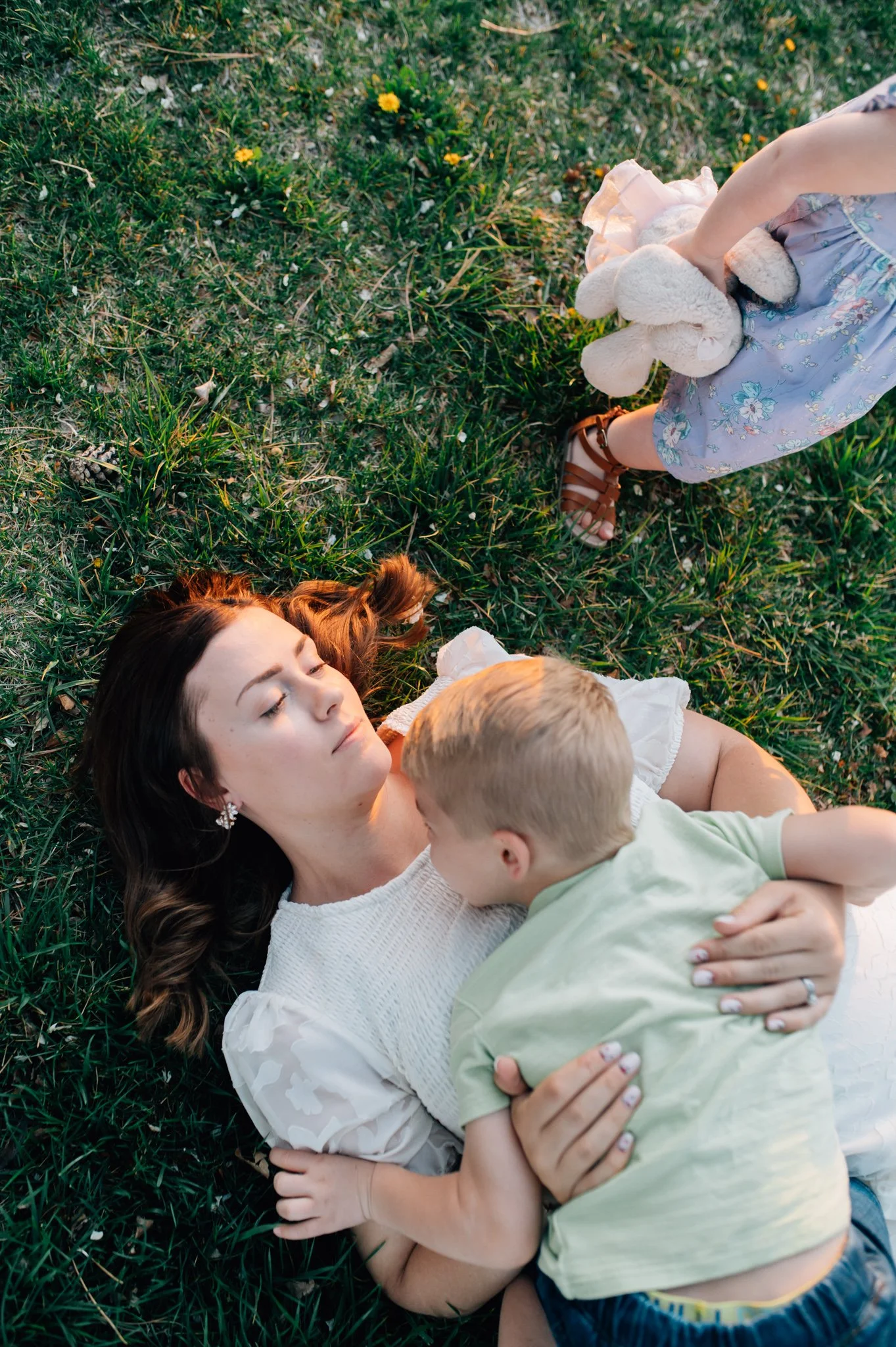 Mom laying in grass holding her son while her daughter holding a stuffed animal walks away