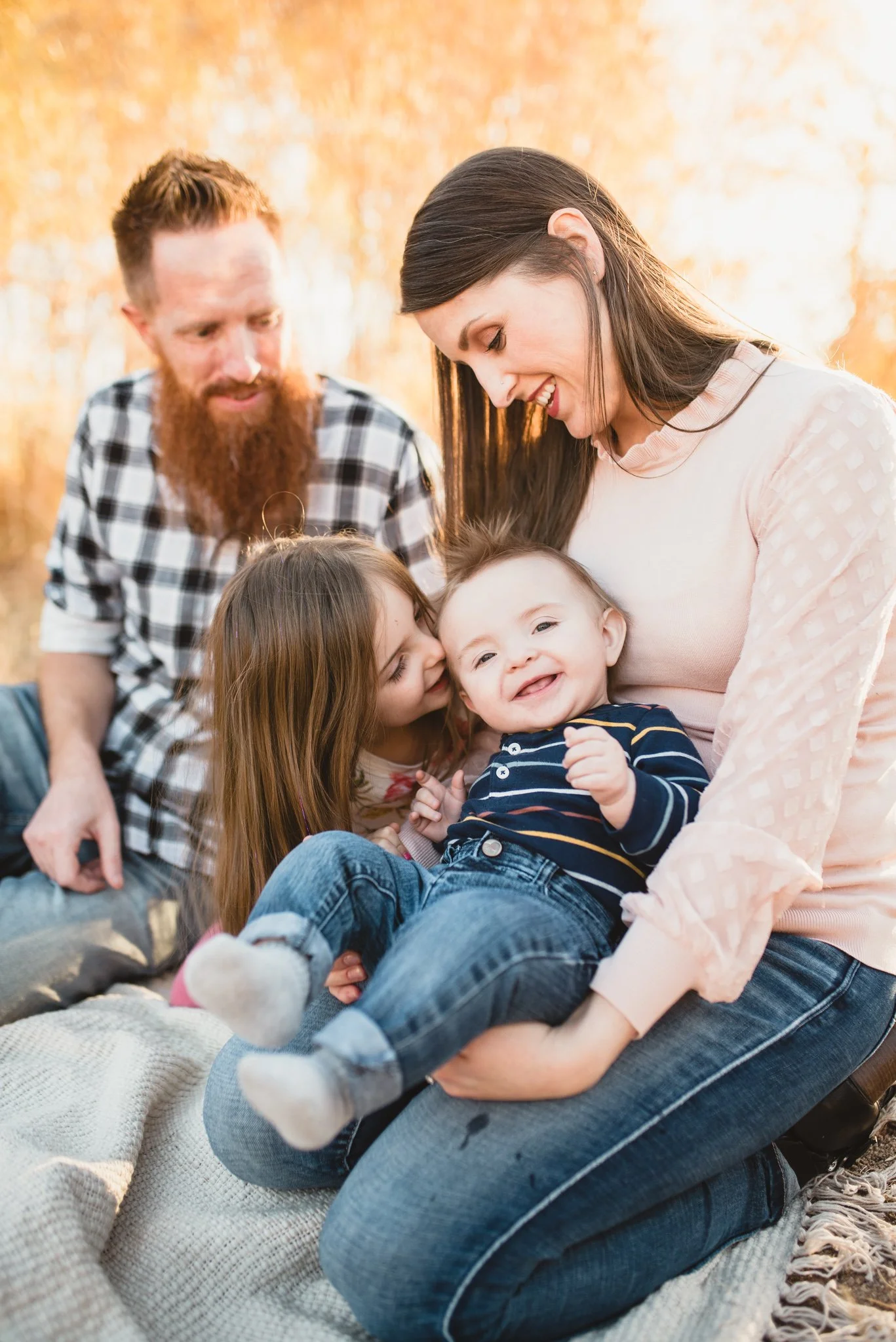 Family of four sitting on blanket, parents look on as older sister kisses baby brother on the cheek