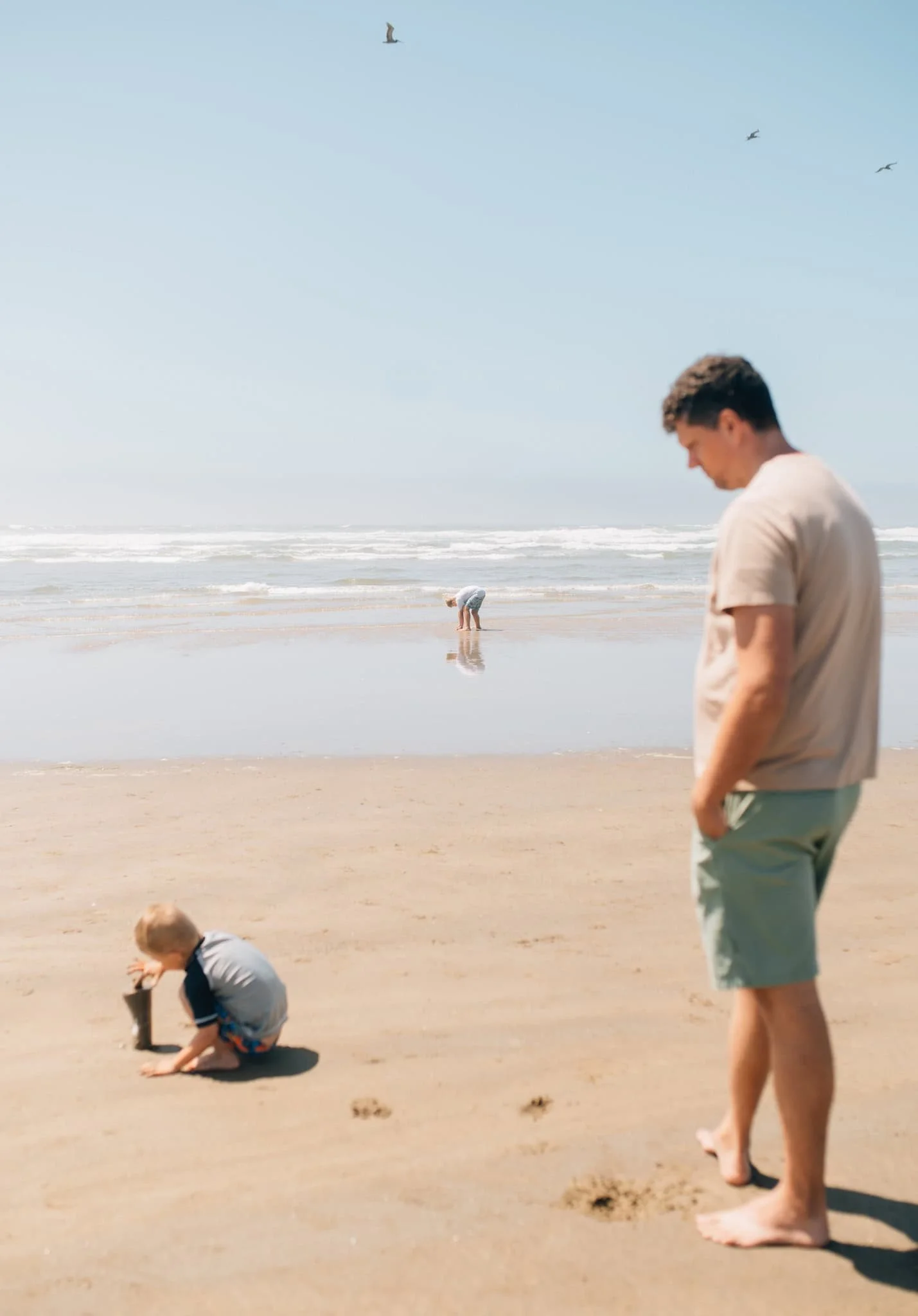 Dad looks on as one son plays in the sand and the other plays in the water