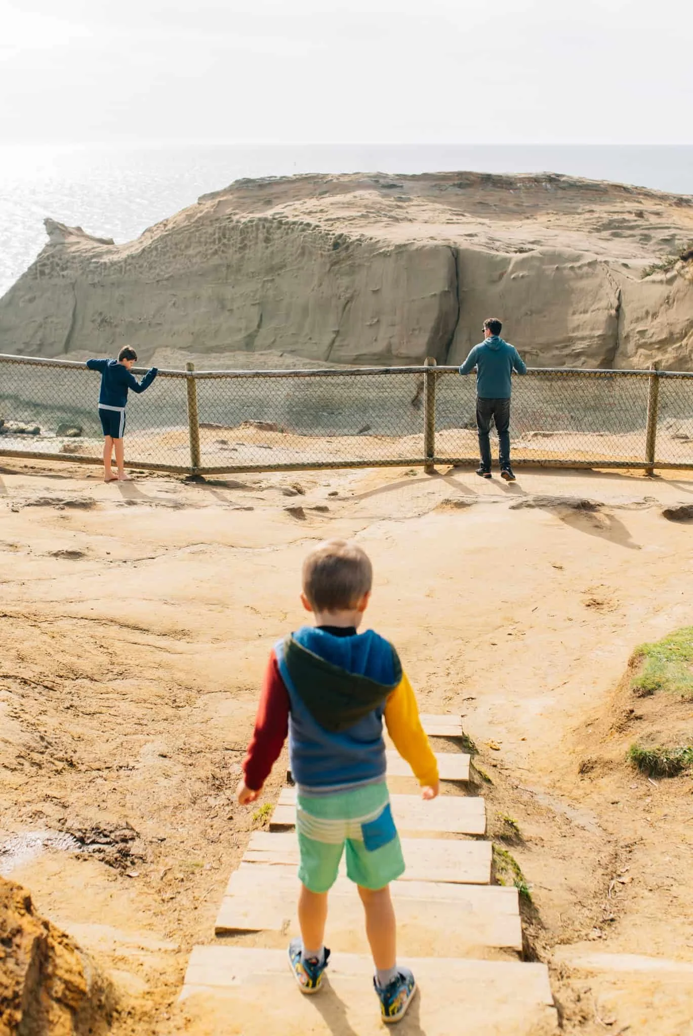 A little boy walks down stairs towards his dad and brother at a gated lookout at Pacific City Beach, OR