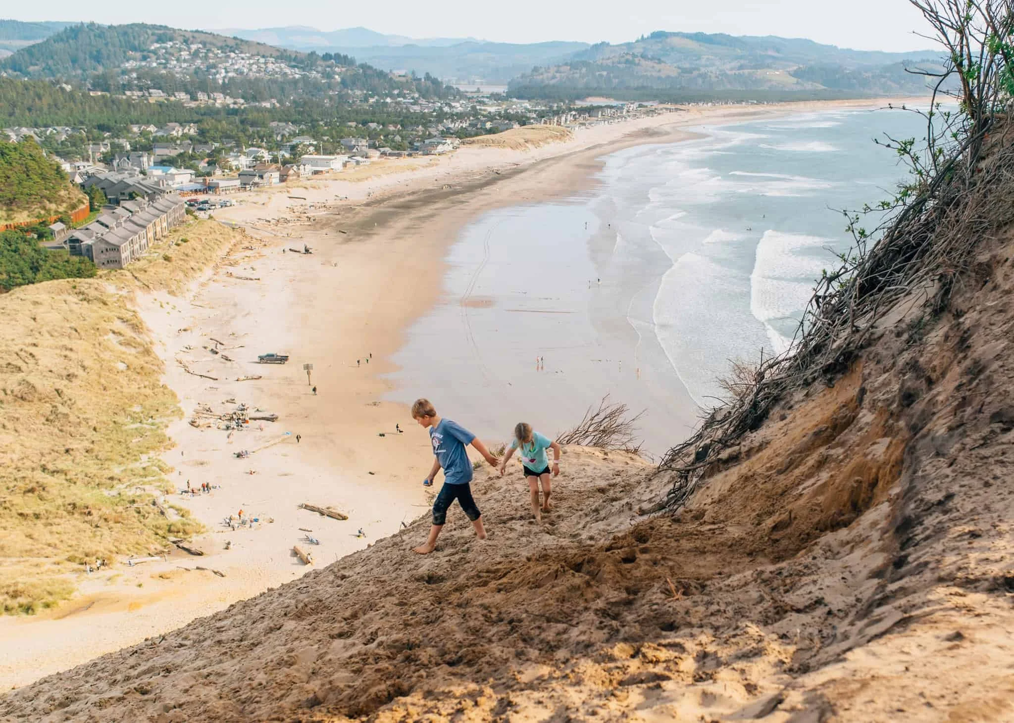 A boy and girl walk along the top of a sand dune with Pacific City Beach far below them