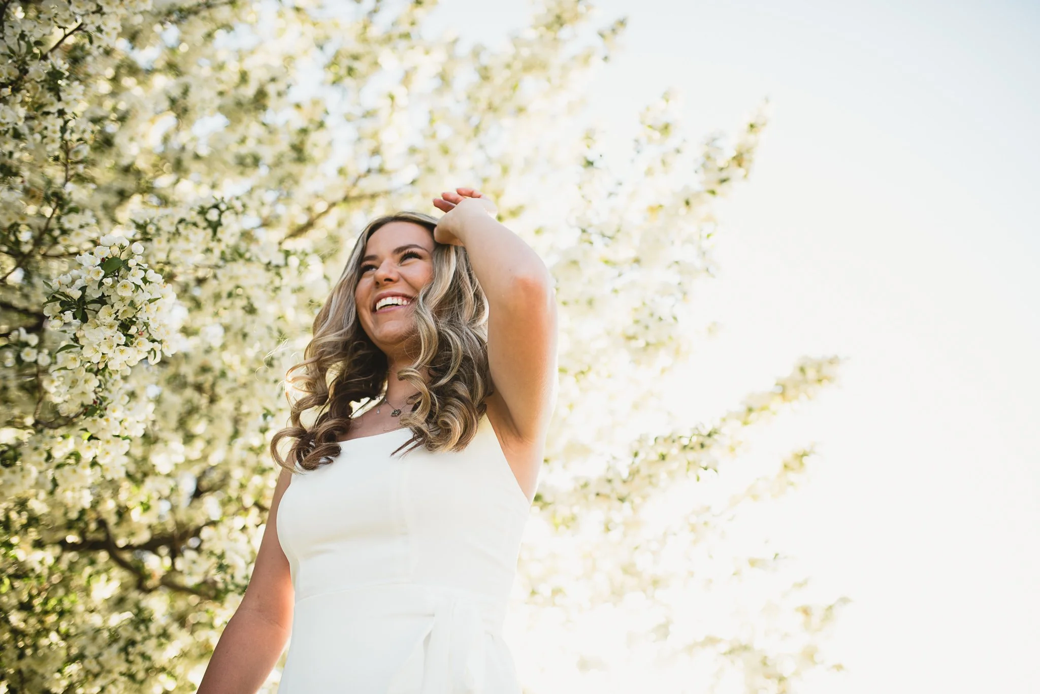 Senior girl in white dress sweeps hair out of face with white blossoming tree behind her