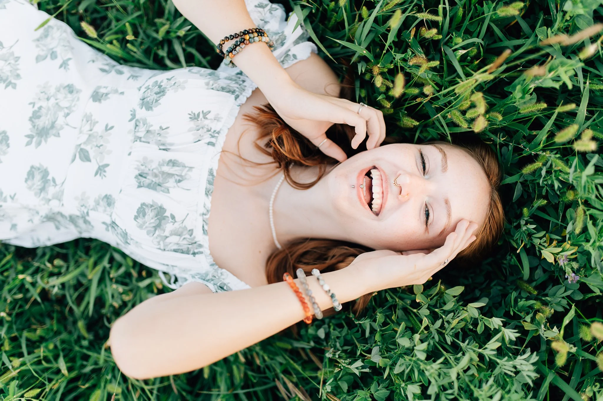 Senior girl in white dress lays in grass and laughs up at camera