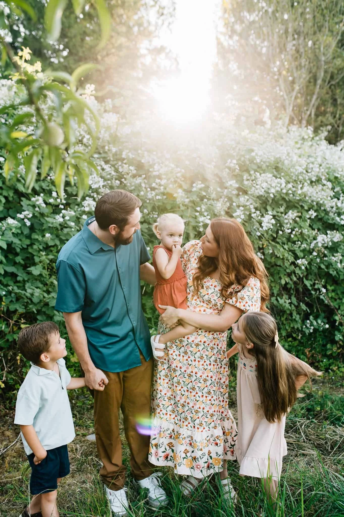 Family with three kids looks at youngest toddler being held by mom with white flowering bushes in the background
