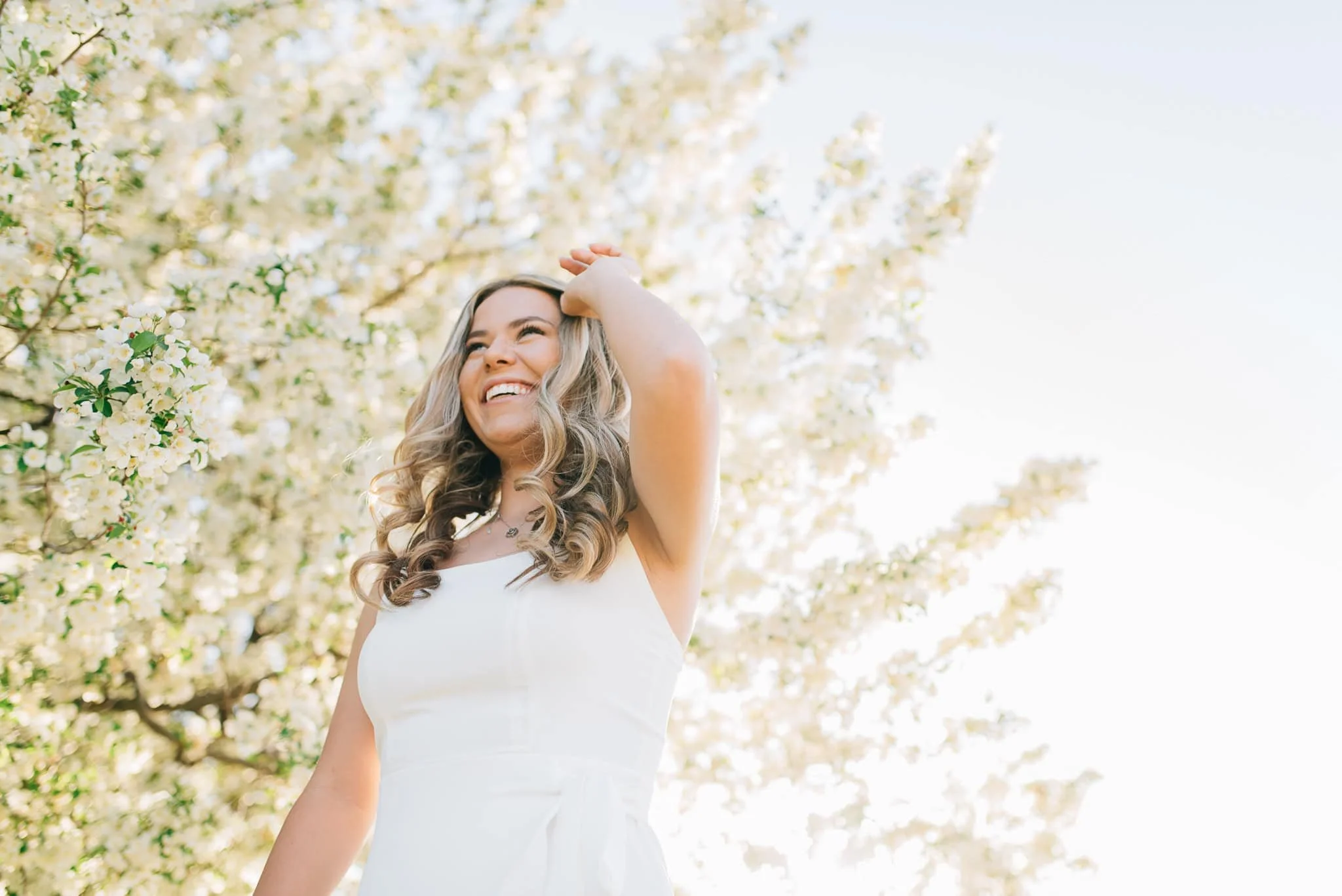 senior girl in white dress runs hand through hair with white flowering tree behind her