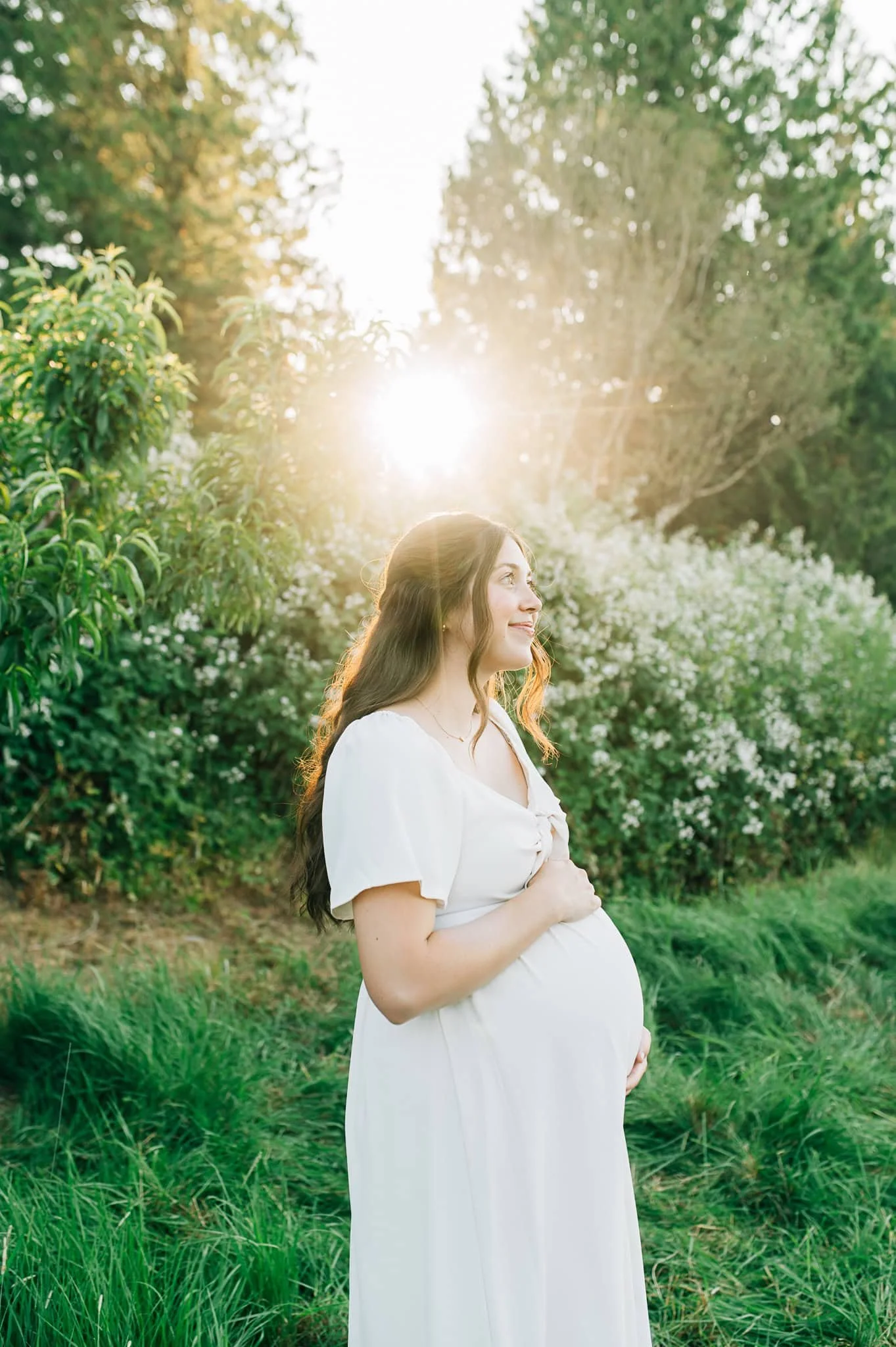 Woman with long hair and white dress holds pregnant belly with sun and white flowery bush behind her