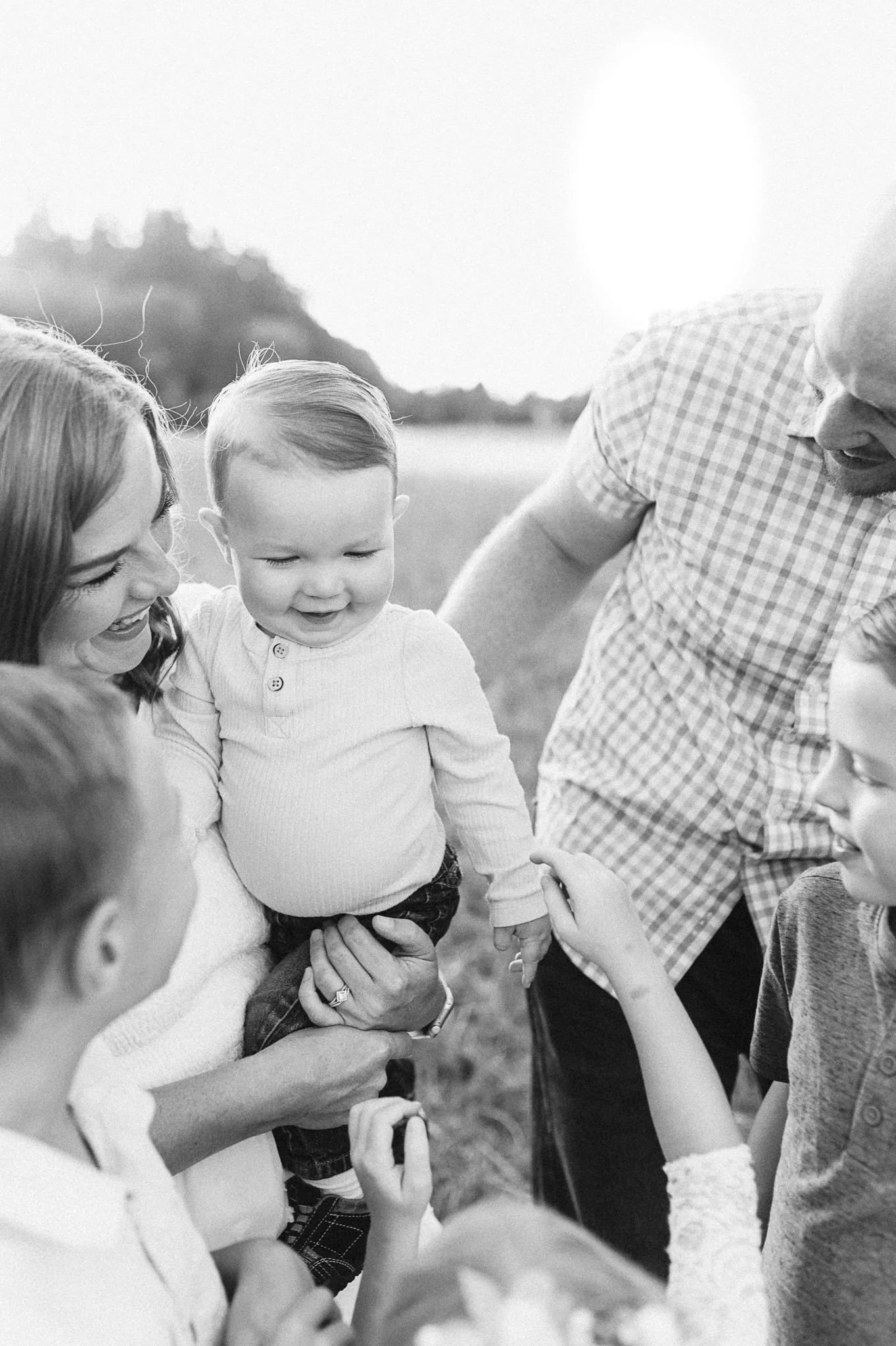Mom holds baby while three other kids and dad all surround and reach for him
