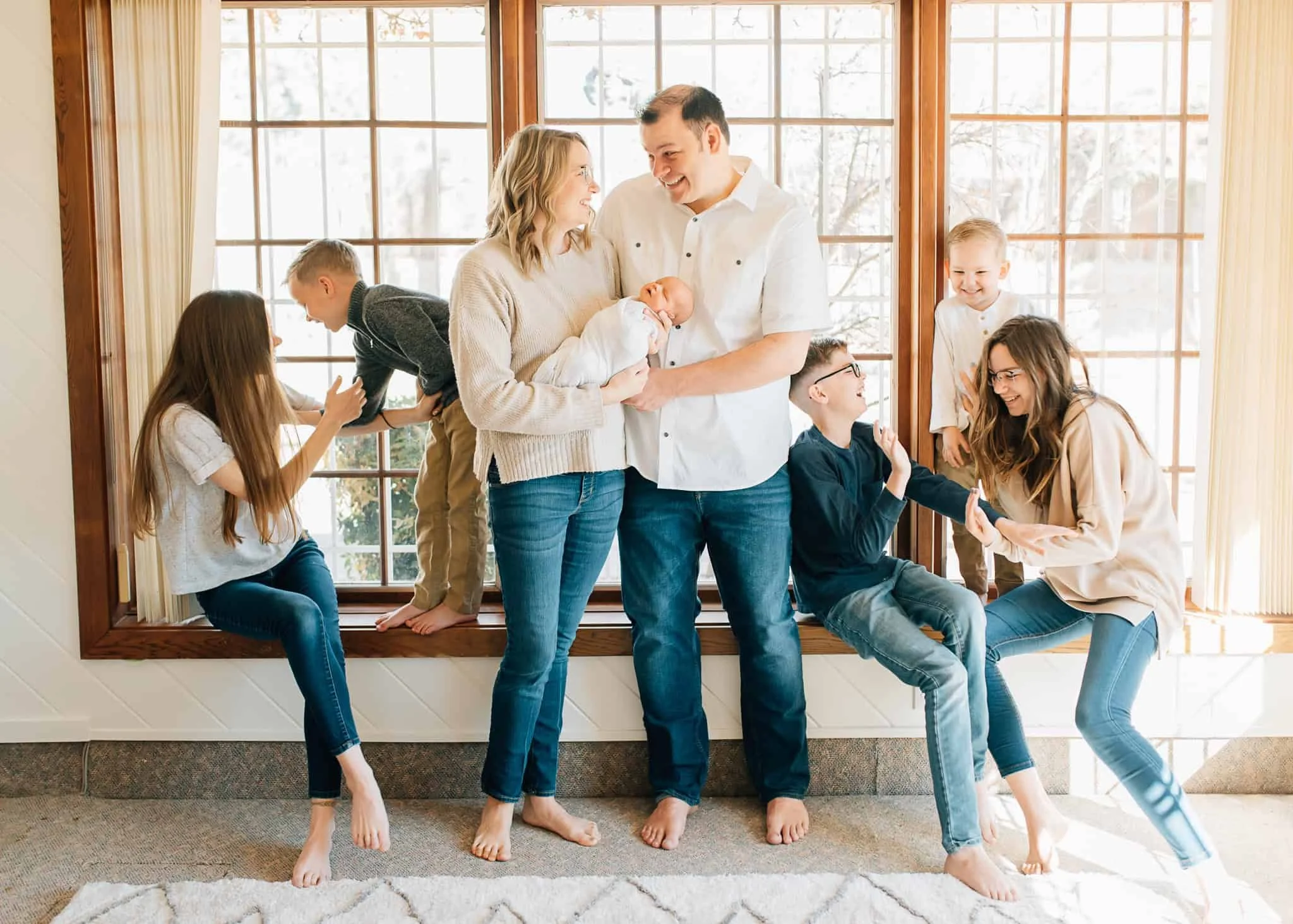 Mom and dad stand together holding newborn while five kids sit on window sill and tickle each other on either side of mom and dad