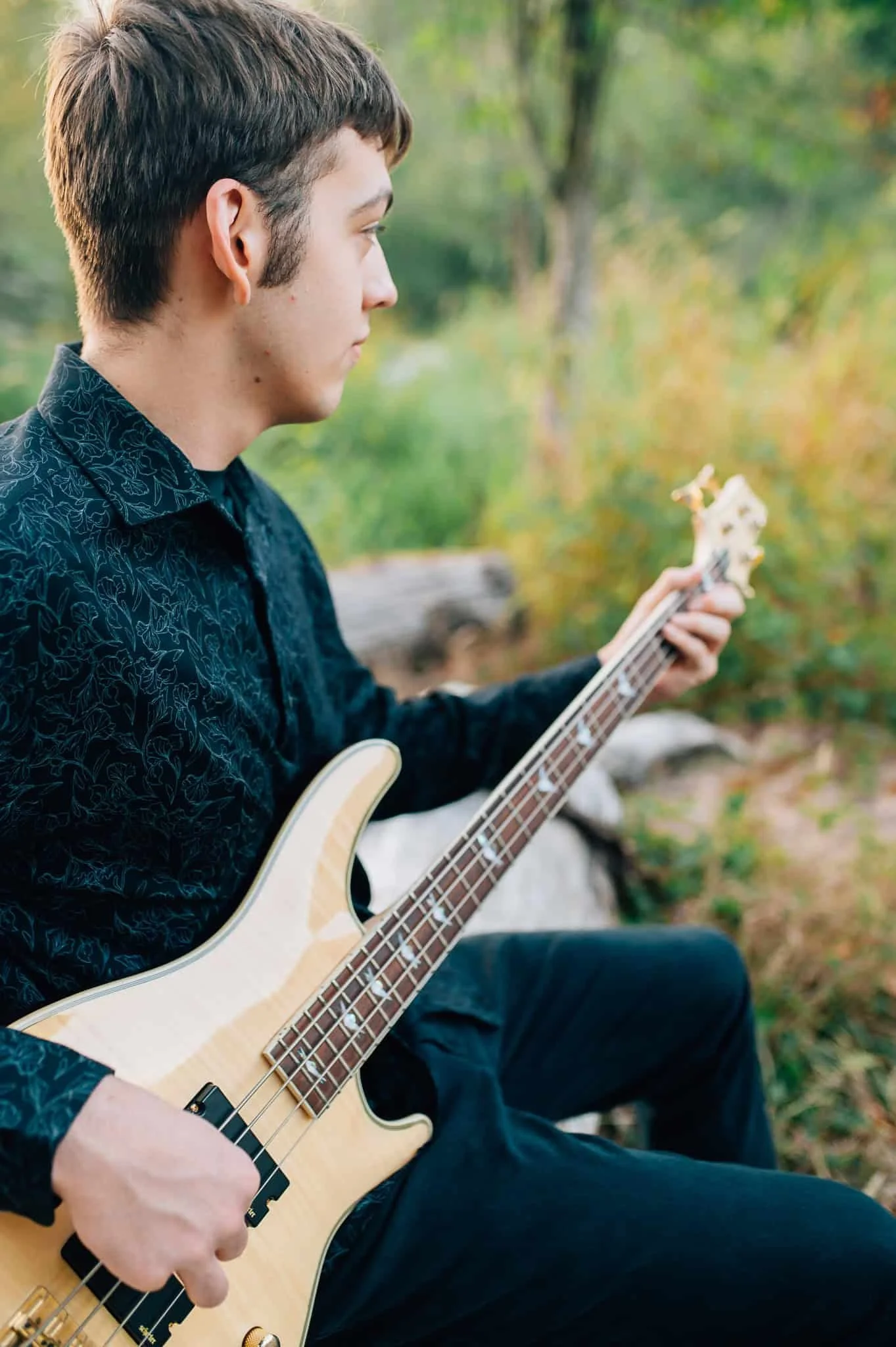 senior boy in black shirt and pants sits on log and plays guitar