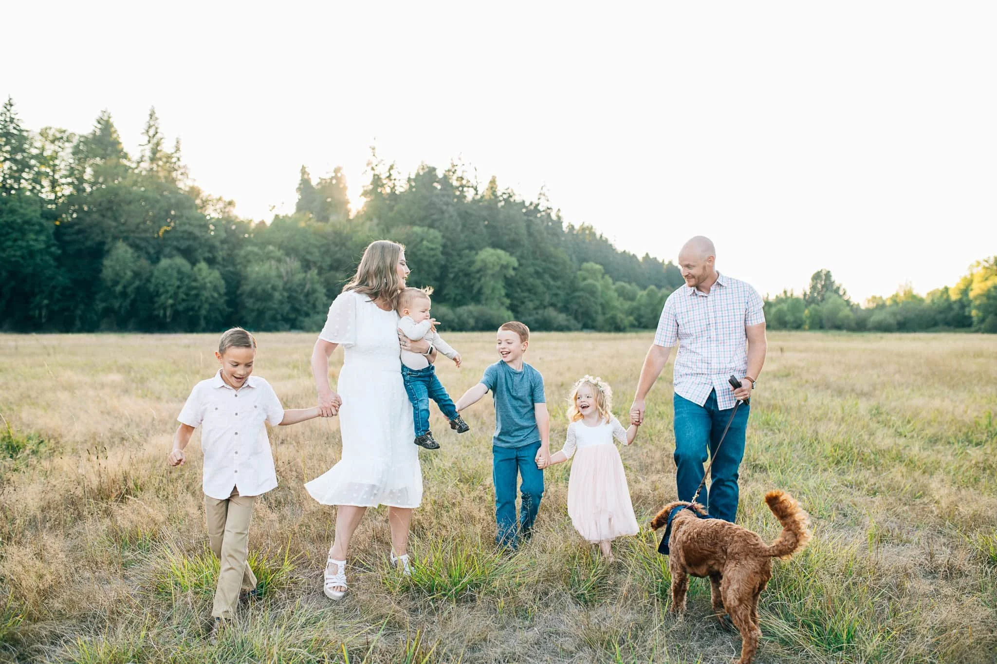 Family with four kids and dog holding hands while walking in a field with trees in the back in Vancouver WA