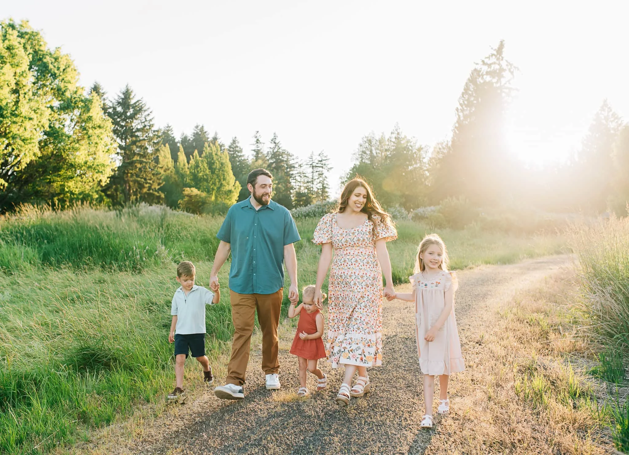 Family with three kids walks down dirt pathway holding hands surrounded by green grass and trees