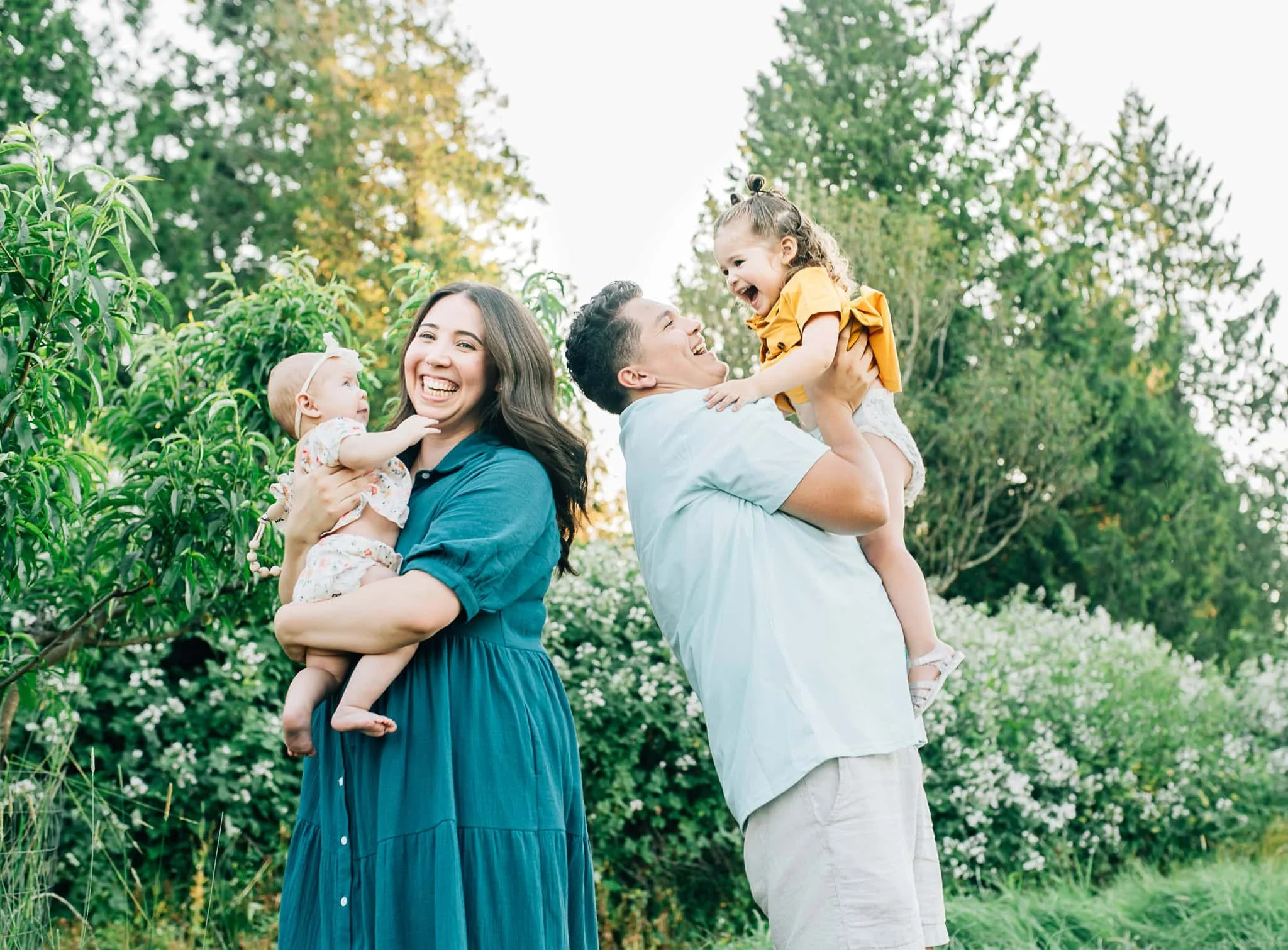 Mom holds baby with back against husband who is holding their toddler while they all laugh