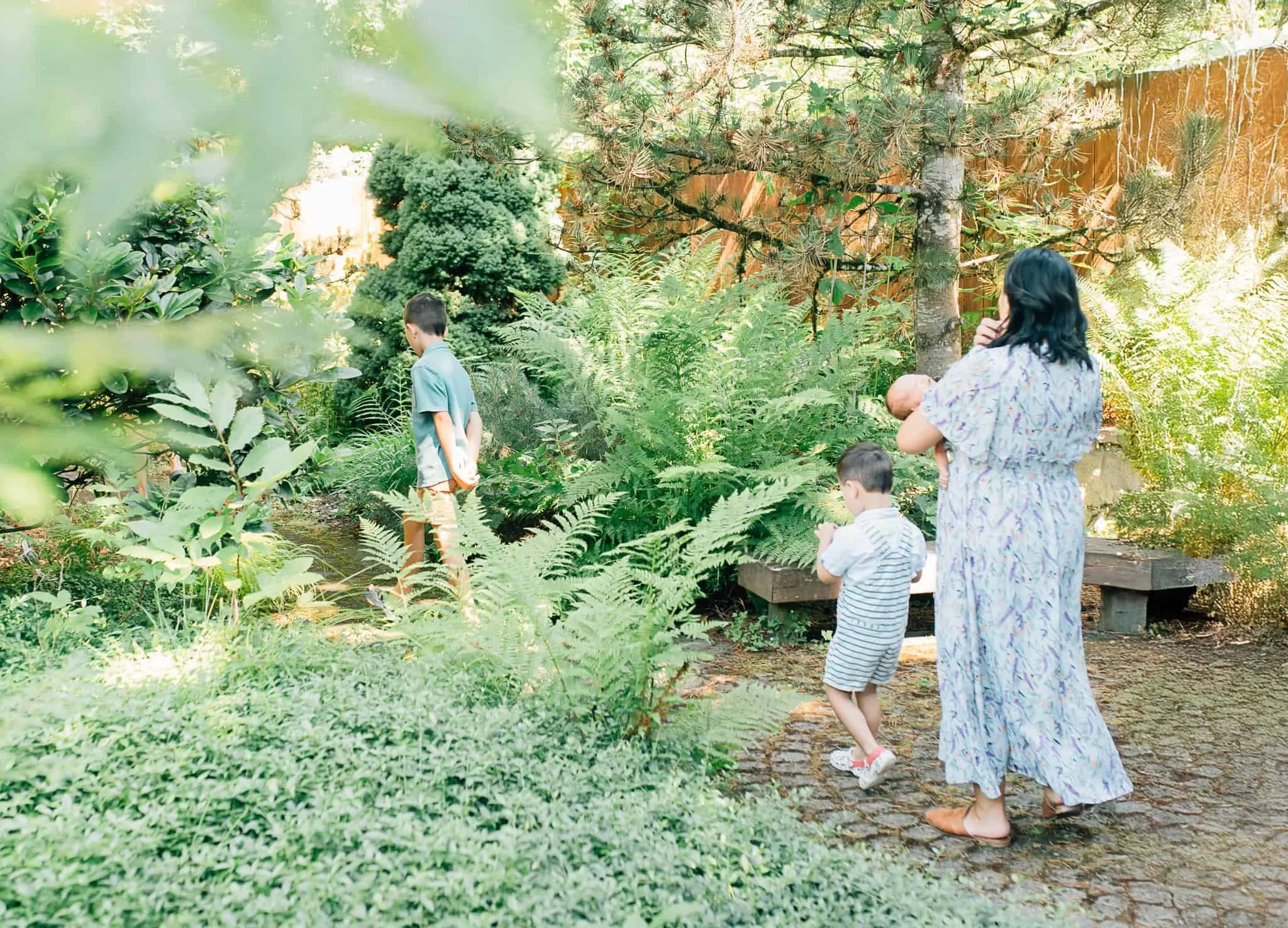 Mom in blue dress carries newborn baby while following her two sons through their backyard garden in Vancouver WA