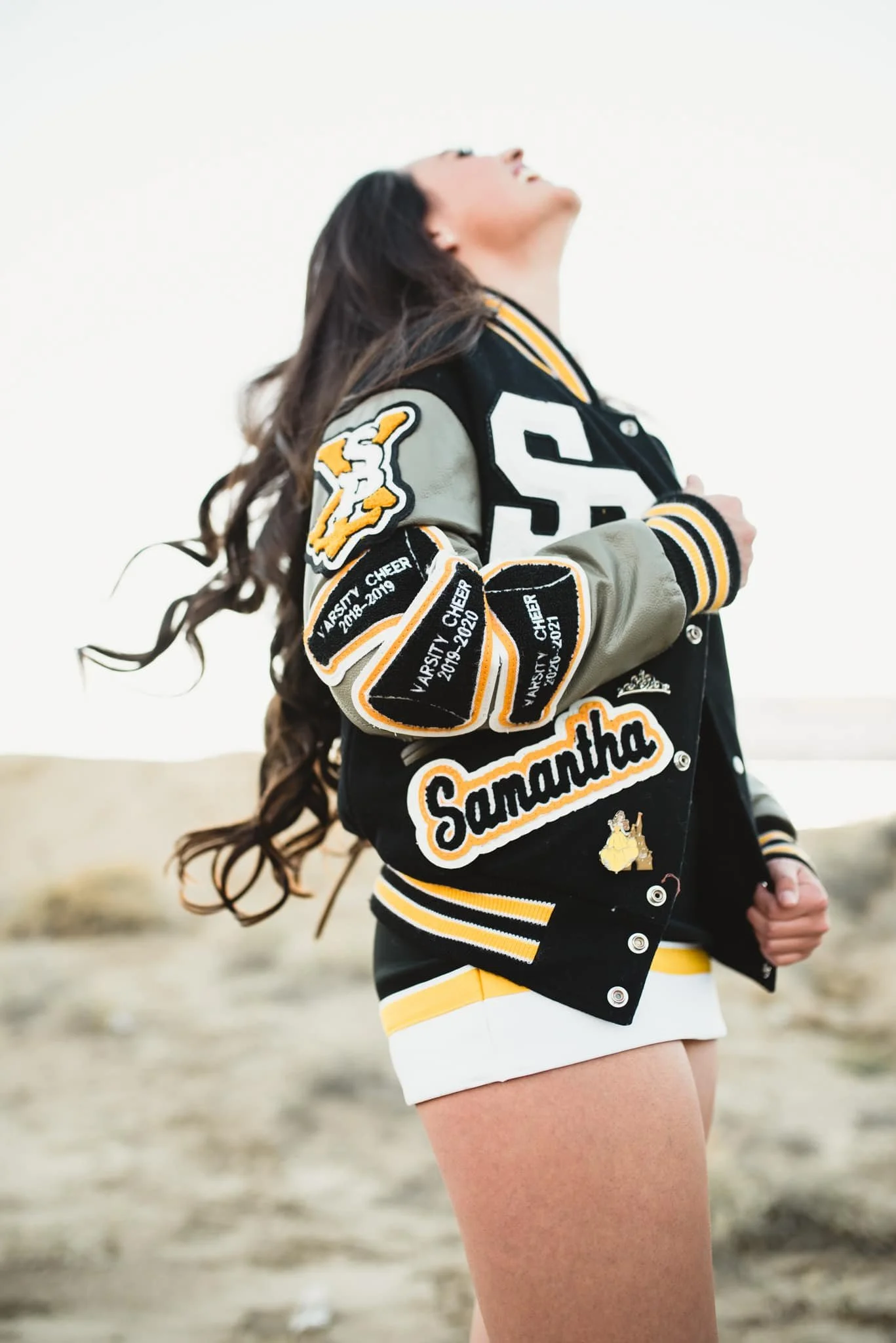 senior girl wearing letterman jacket throws head back and laughs at sky