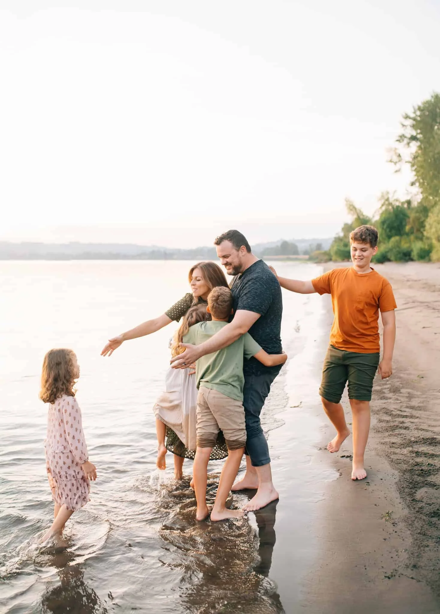 Family with four kids stands on the shoreline wading into the water as they hug two kids while reaching for the other two kids