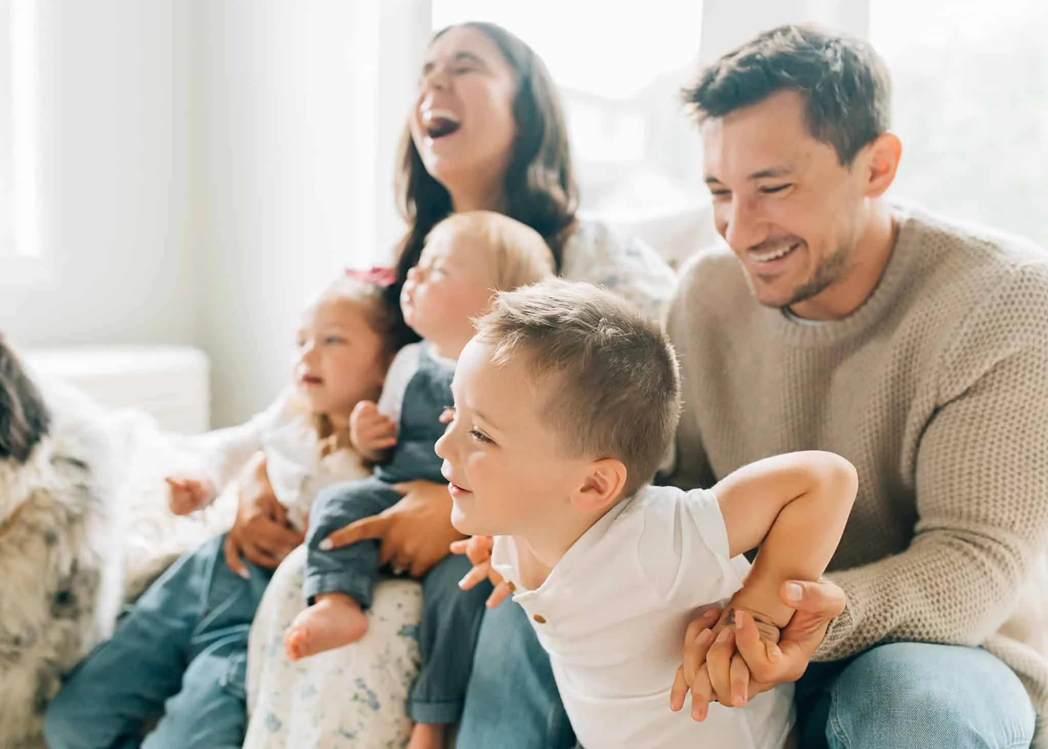 Family with three young kids sits with kids on laps while mom throws head back and laughs