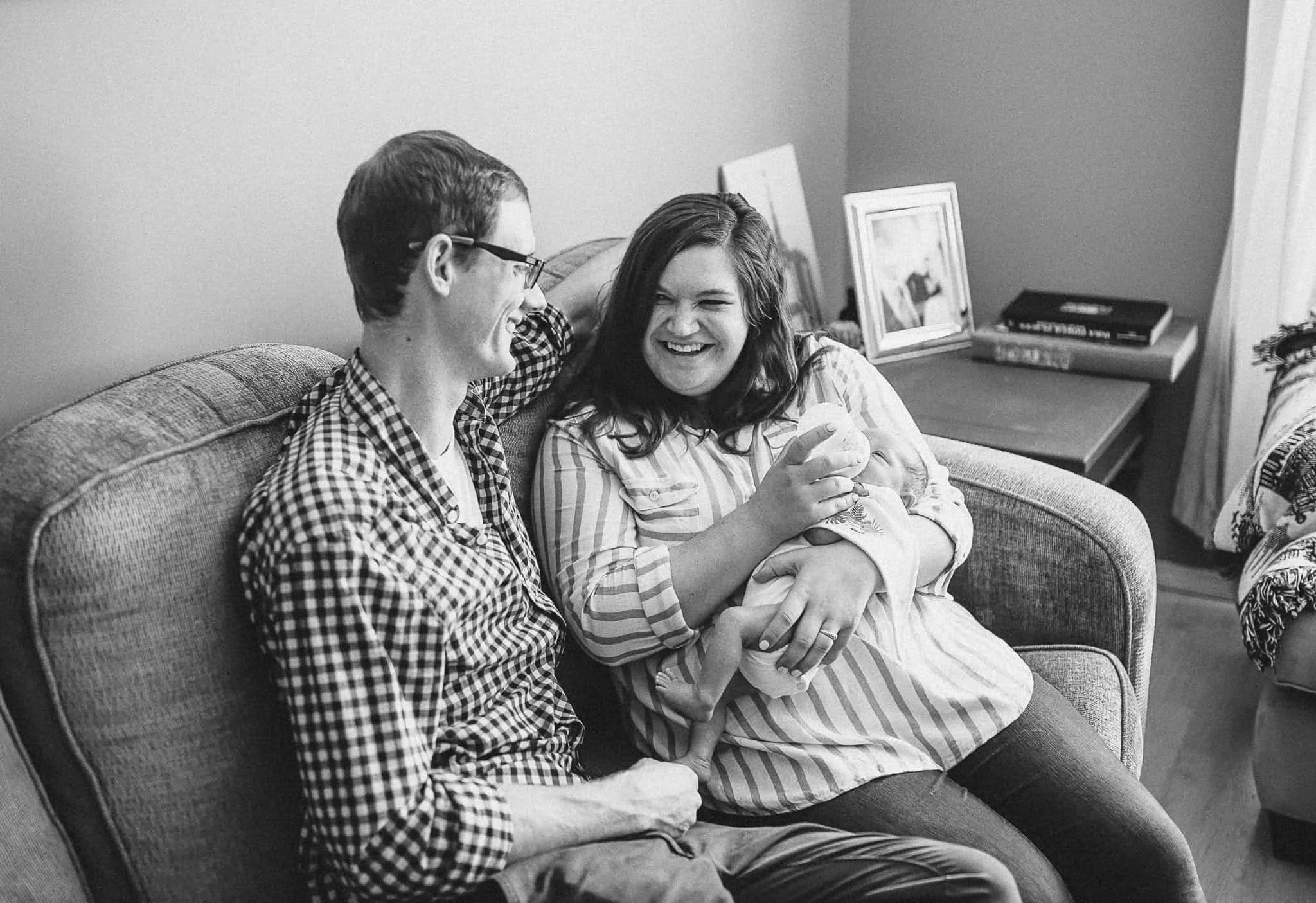 husband and wife sitting on couch and laughing together while mom feeds bottle to newborn baby