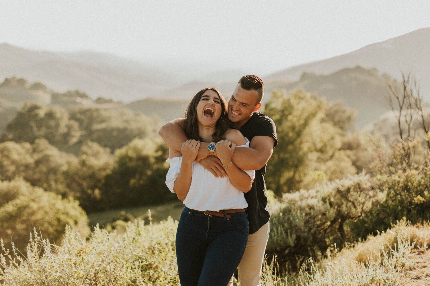 couple laughs together with their arms wrapped around one another overlooking the hills of paso robles during their engagement photos. san luis obispo proposal photographer