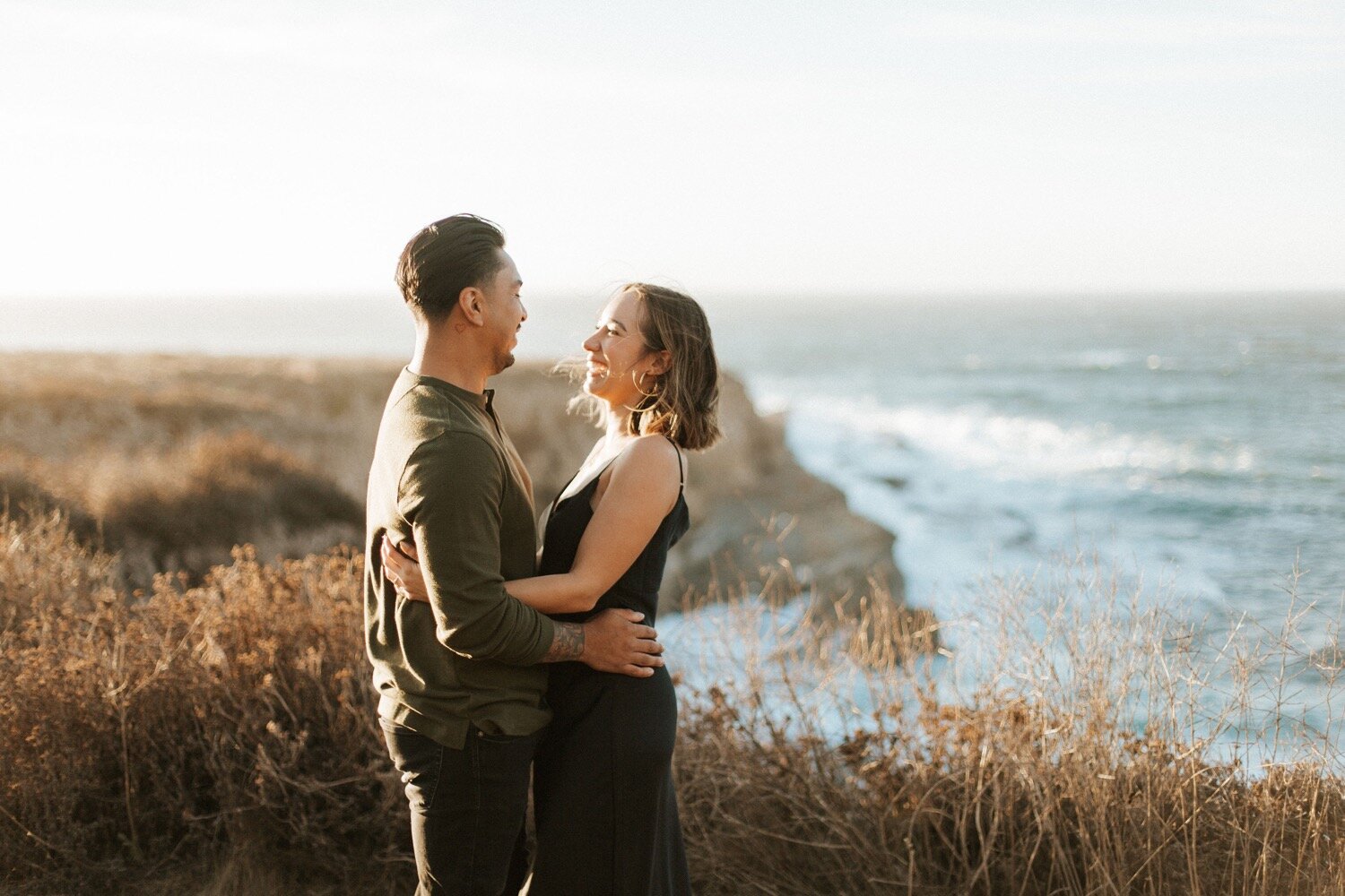  couple holds each other and laughs as the sun sets behind them on the bluff trail at montana de oro state park. couple’s photoshoot captured by slo wedding photography duo poppy and vine 