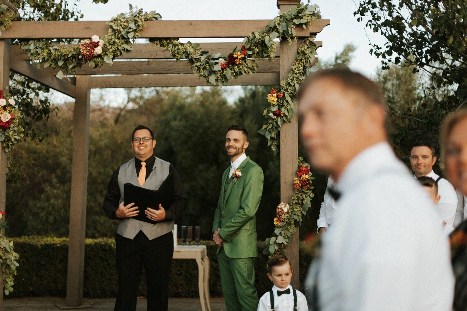 groom, dressed in a green suit, smiles as he sees his bride for the first time walking down the aisle at their dana powers house wedding, captured by slo wedding photographers poppy and vine 
