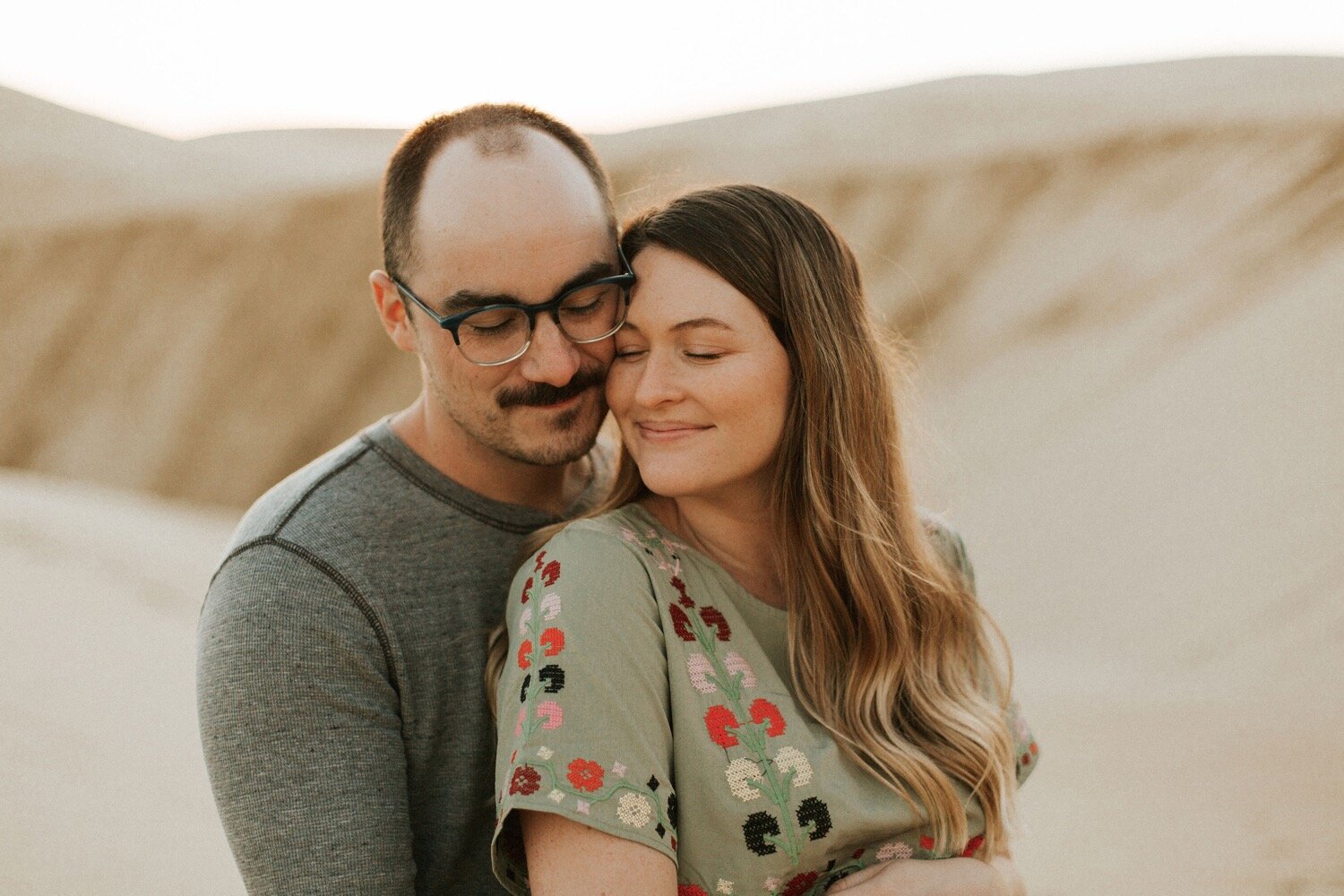  husband and wife snuggle up to each other as the sun sets over the oceano dunes in pismo beach california, photographed by california couple photographer poppy and vine 