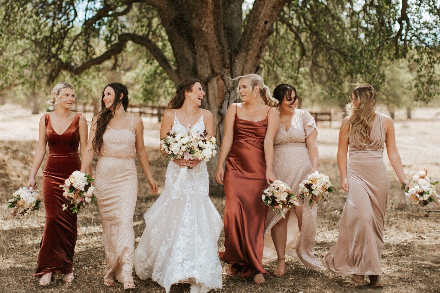  bridesmaids wearing mismatched rust and neutral colored bridesmaids dresses walk together with the bride at her boho ranch wedding in new cuyama, captured by slo wedding photographer poppy and vine 