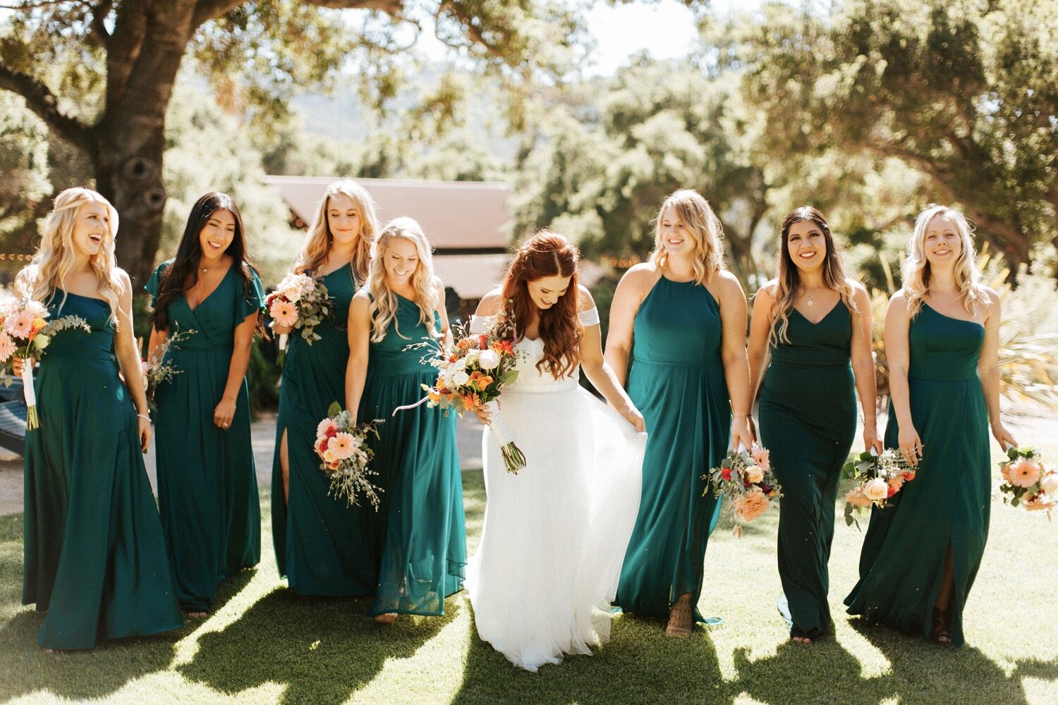  bride, wearing wtoo by watters, and bridesmaids, wearing jewel toned floor length azazie dresses, walk together at a sunny spring afternoon wedding at the loomis family barn in arroyo grande, california, captured by san luis obispo wedding photograp