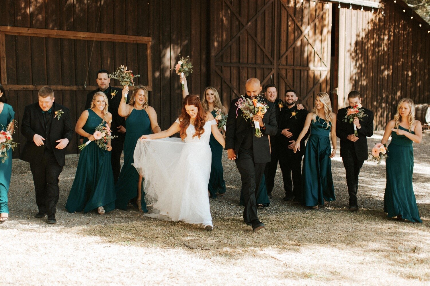  bridesmaids and groomsmen, dressed in jewel toned outfits, walk with the bride and groom, toting colorful wildflower bouquets, during a wedding at the loomis family barn in arroyo grande, california, captured by slo wedding photographer poppy and vi