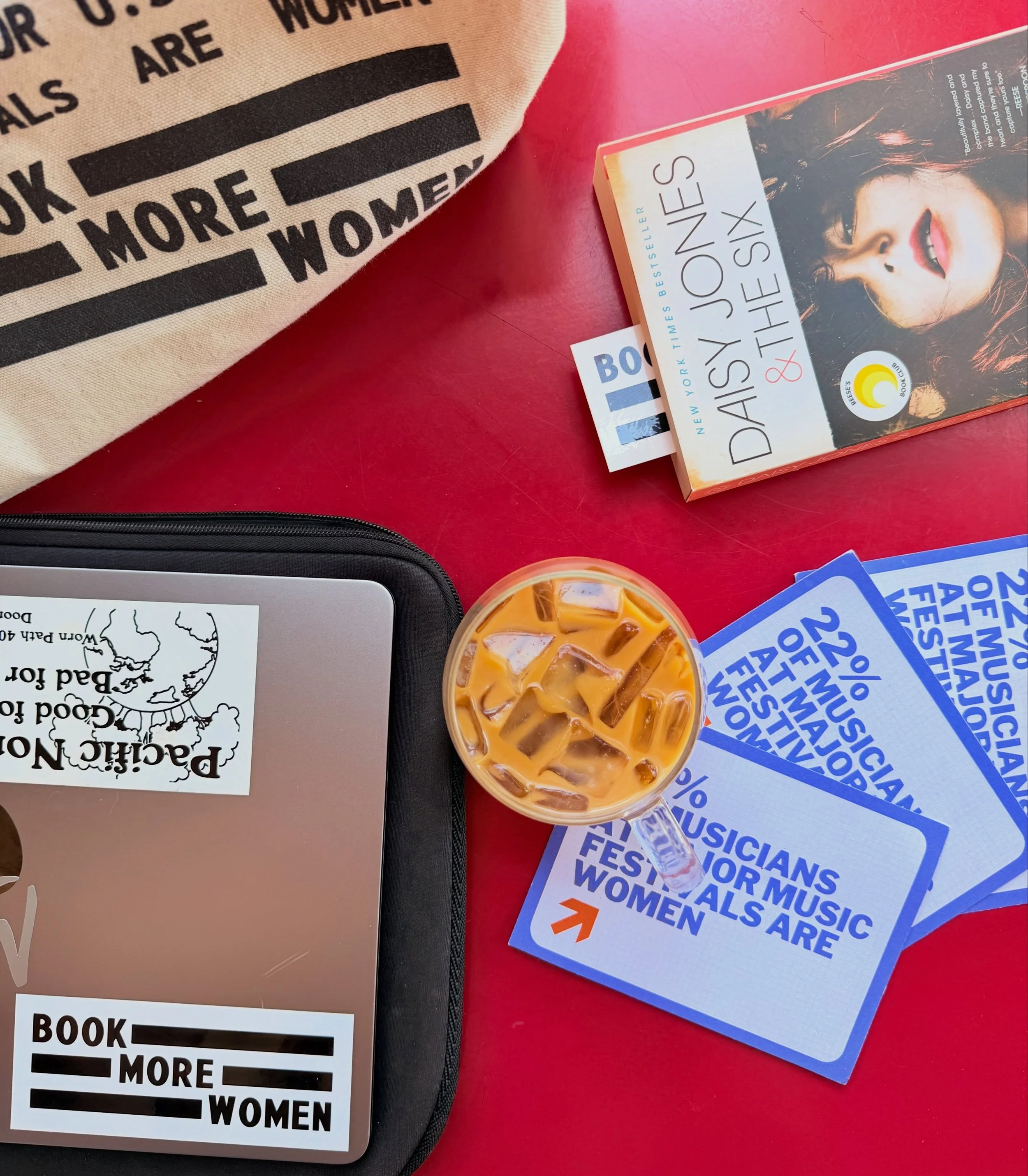 A desktop with Book More Women stickers and tote bag