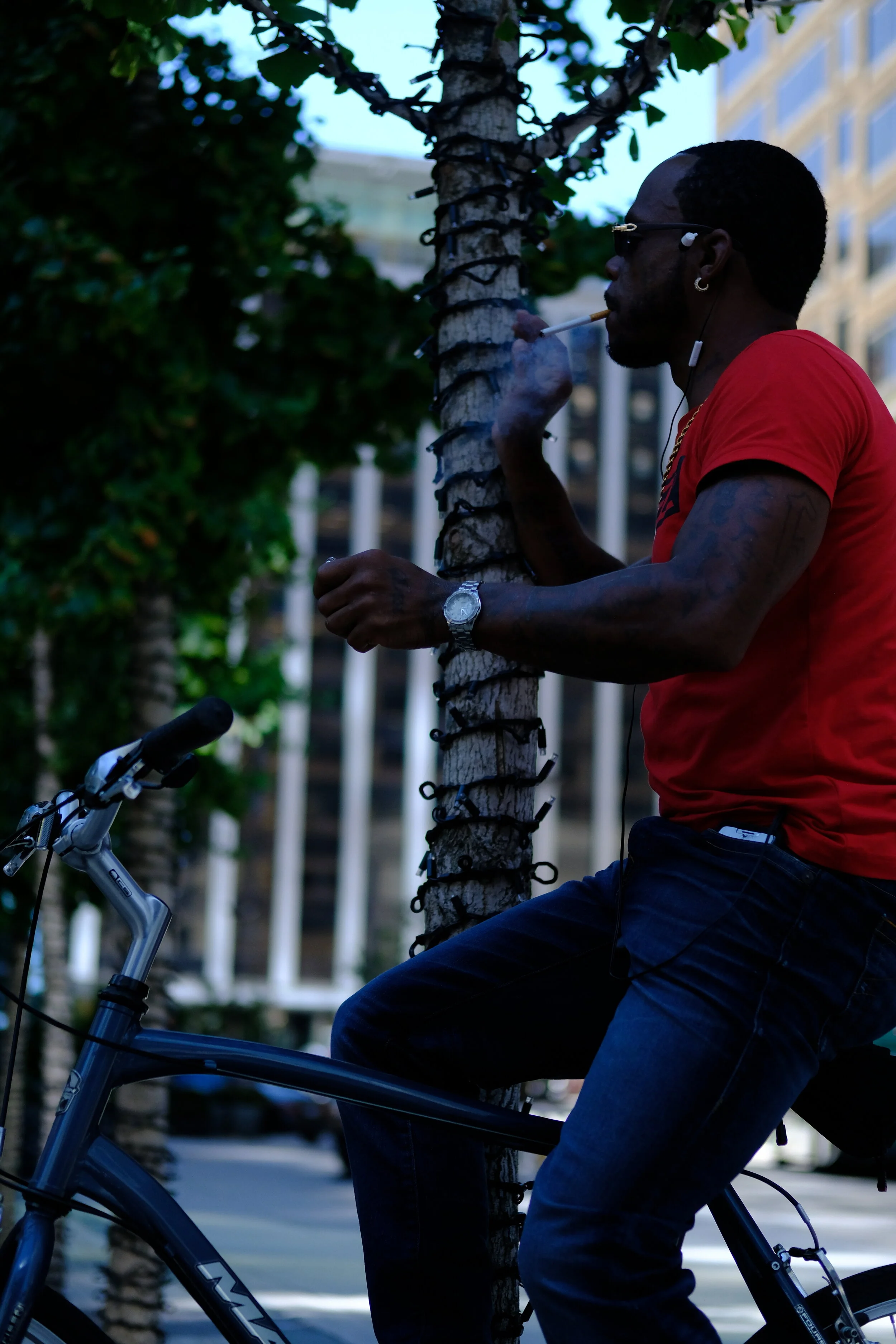  They lean in repose against a tree, precarious on a halted bicycle, indulging a break from their journey toward some unknown but unhurried goal. Smoking cigarettes bummed from the tie-and-suit folk who now and then escape into the loud air of the ci