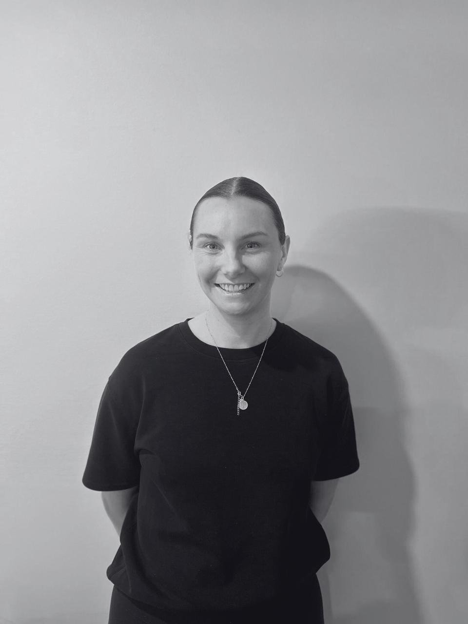 Black and white photo of a woman smiling, wearing a black t-shirt, with her hair slicked back, standing against a plain wall.