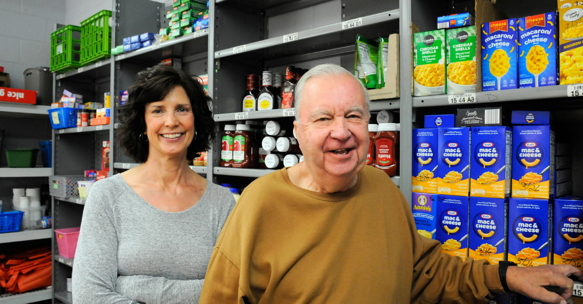 A woman with dark hair and an elderly man stand in front of pantry shelves stocked with food.