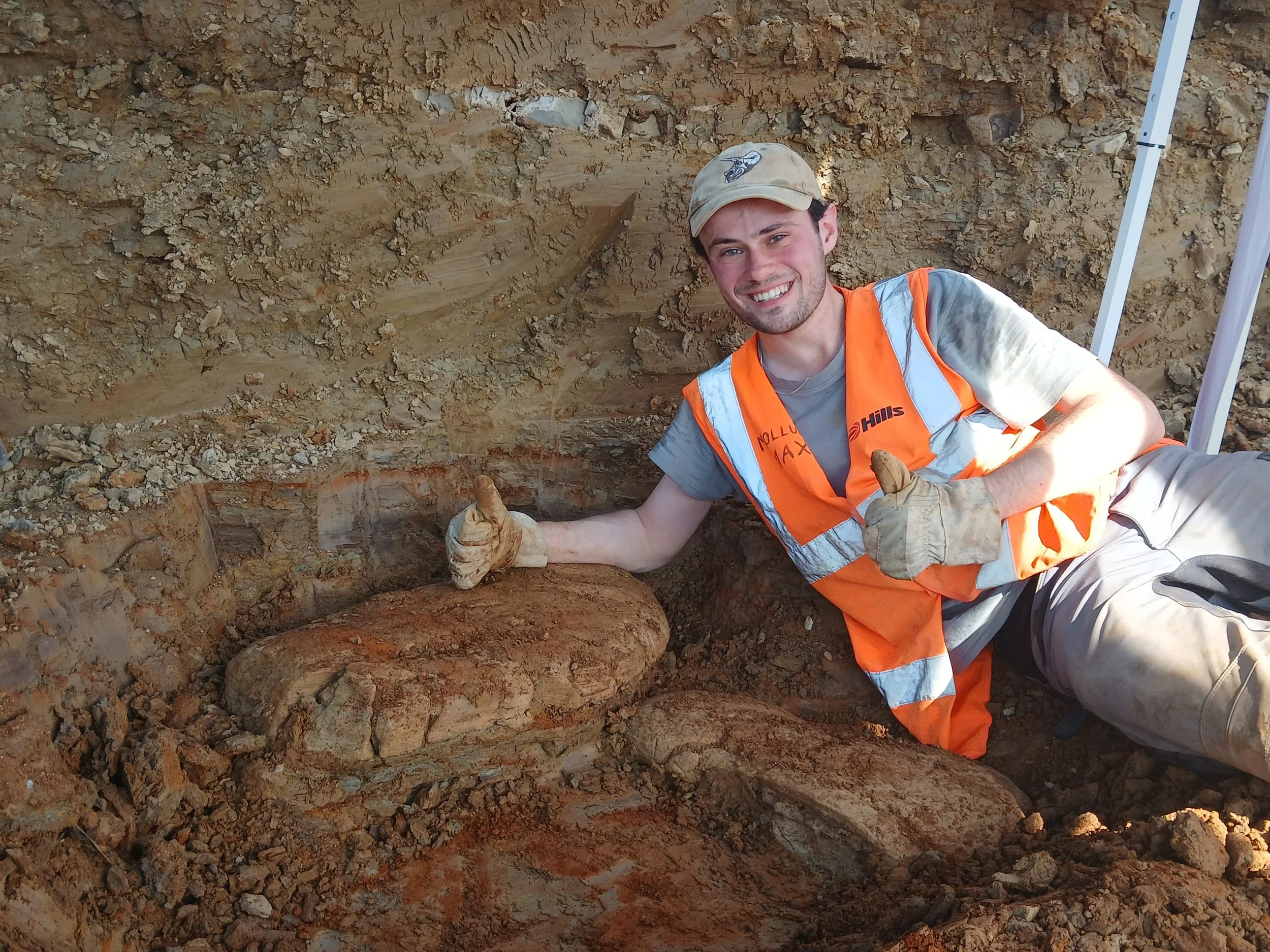 Person wearing high vis lying down doing a thumbs up sign next to a large rock lying in the dirt