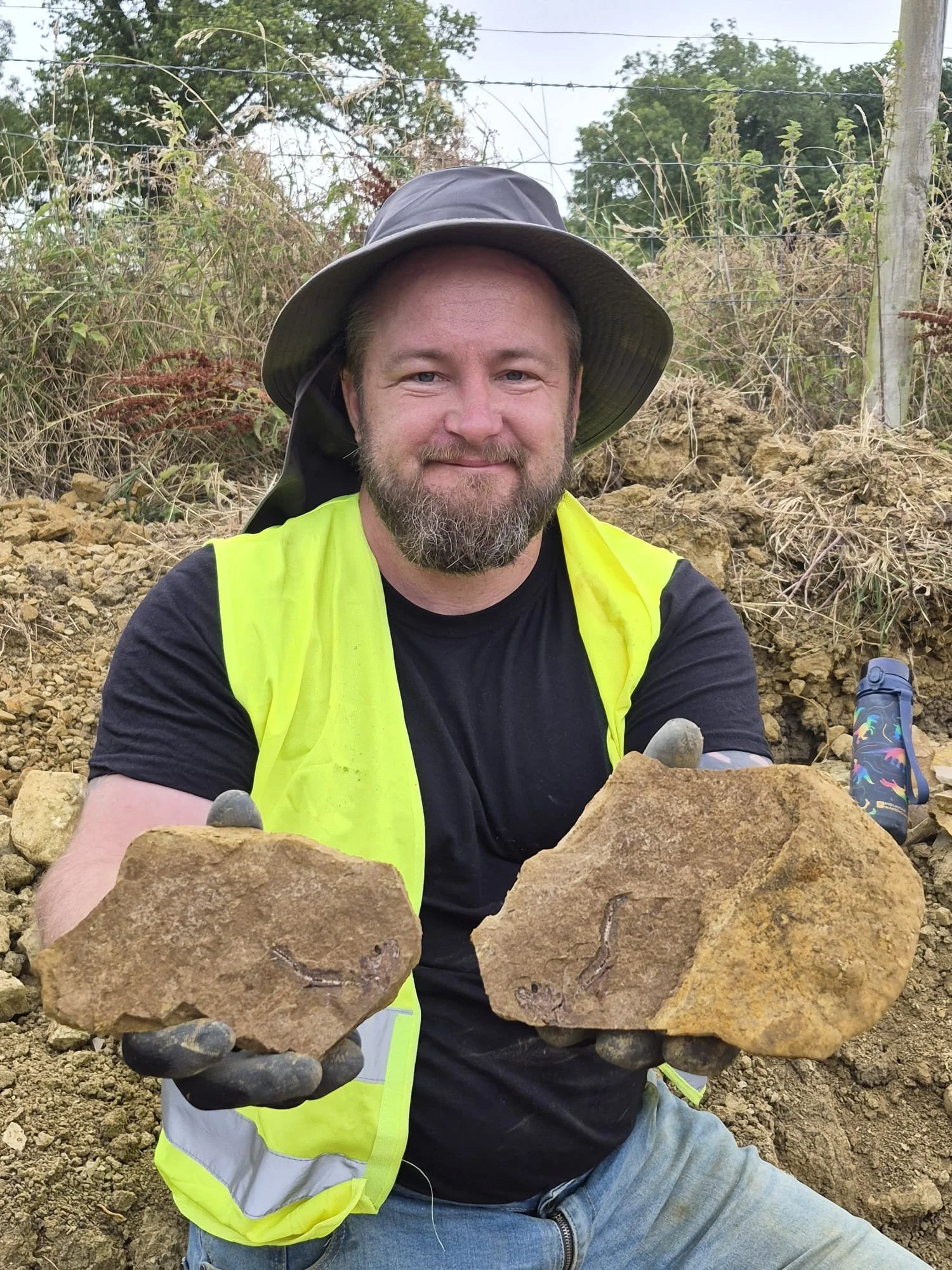 Man in hat and high vis holds up two rocks containing fish fossils