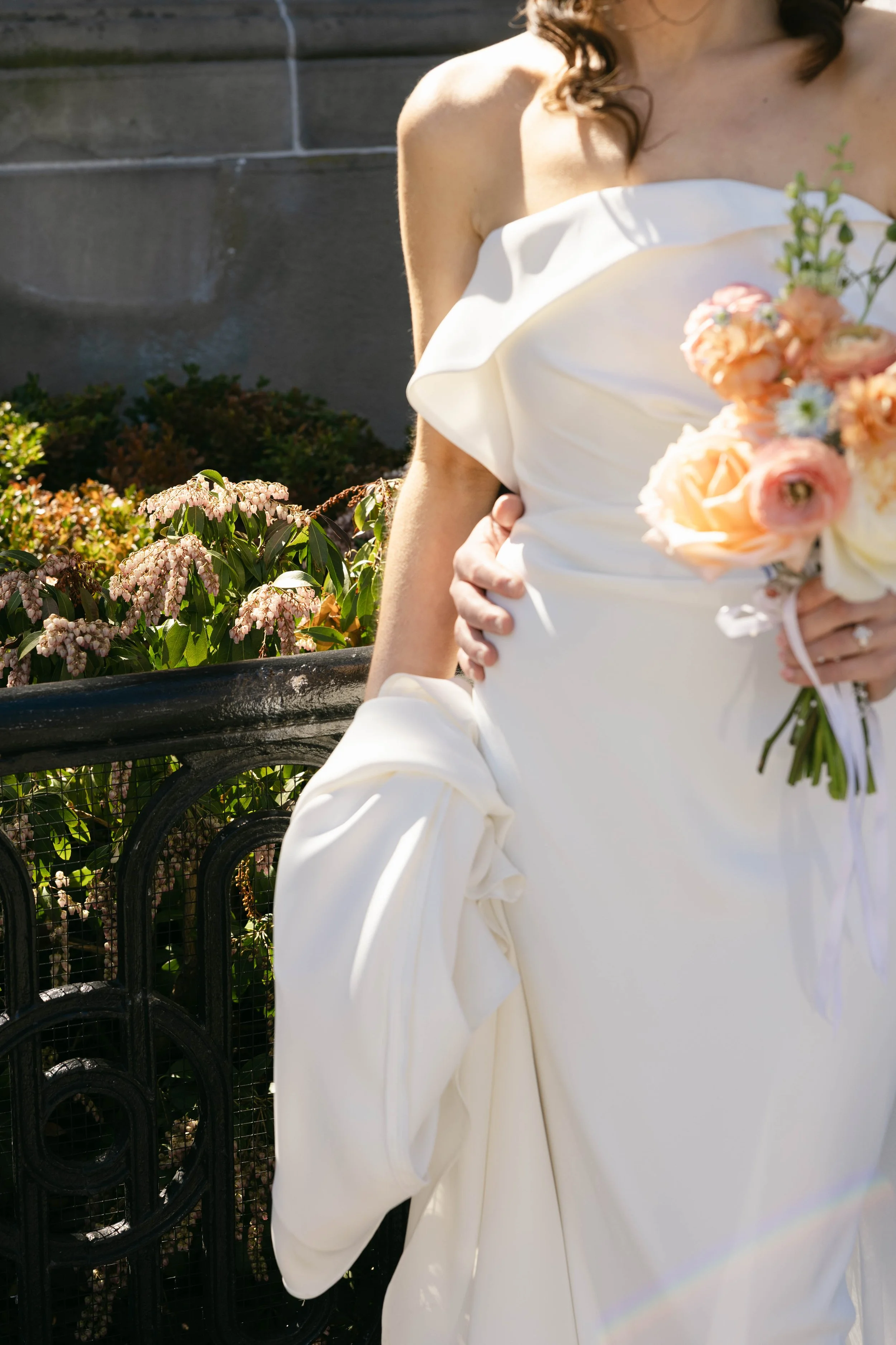 A bride holding a bouquet of pink and white flowers, dressed in a white wedding gown with a one-shoulder ruffle detail, standing outdoors near a black metal fence and green shrubbery with pink flowers.