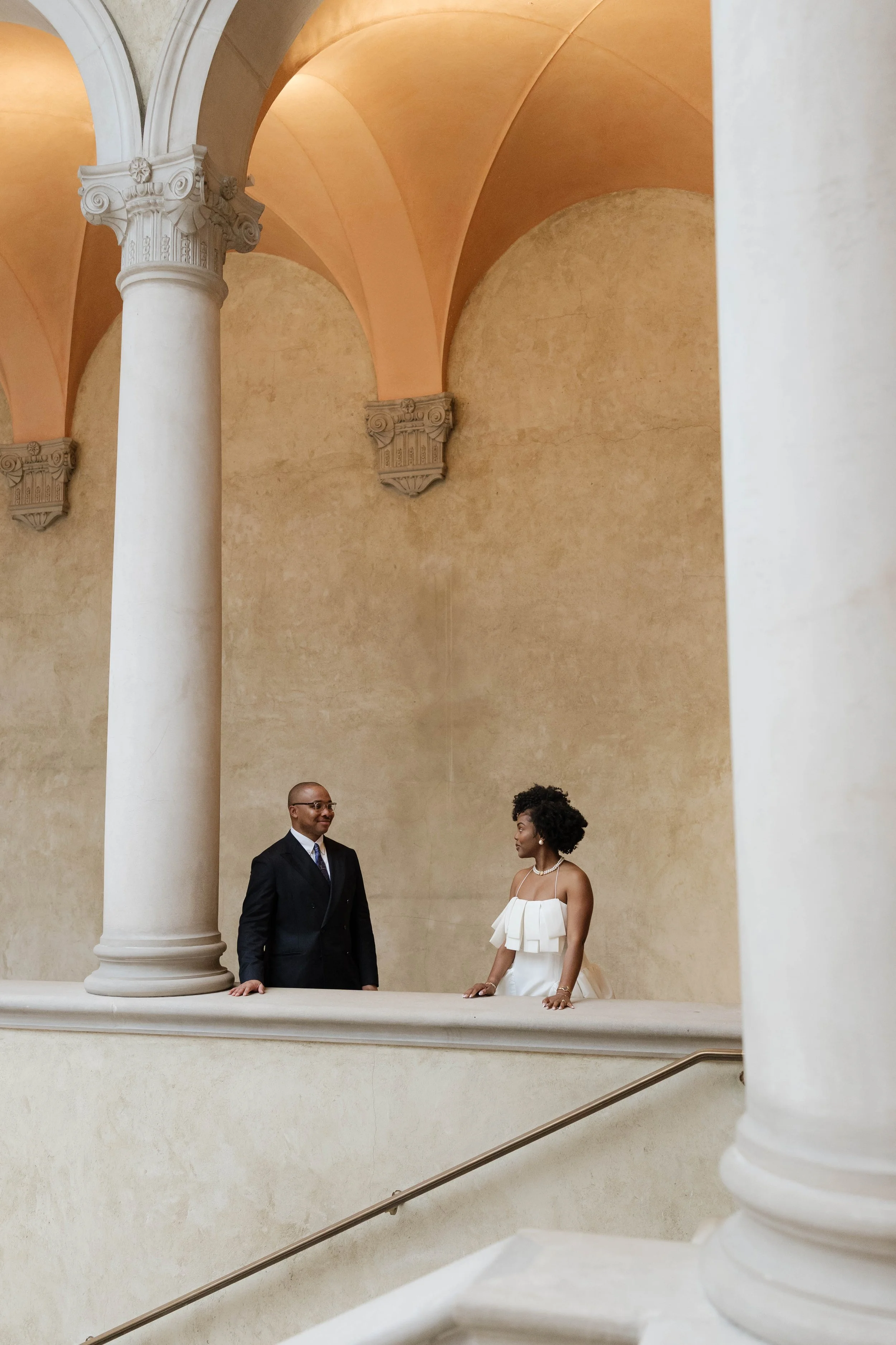 A couple standing on a balcony in a grand, classical-style building with tall columns and arches. The man is wearing a dark suit and glasses, and the woman is in a white dress with ruffled sleeves and pearls. They are looking at each other in a forma