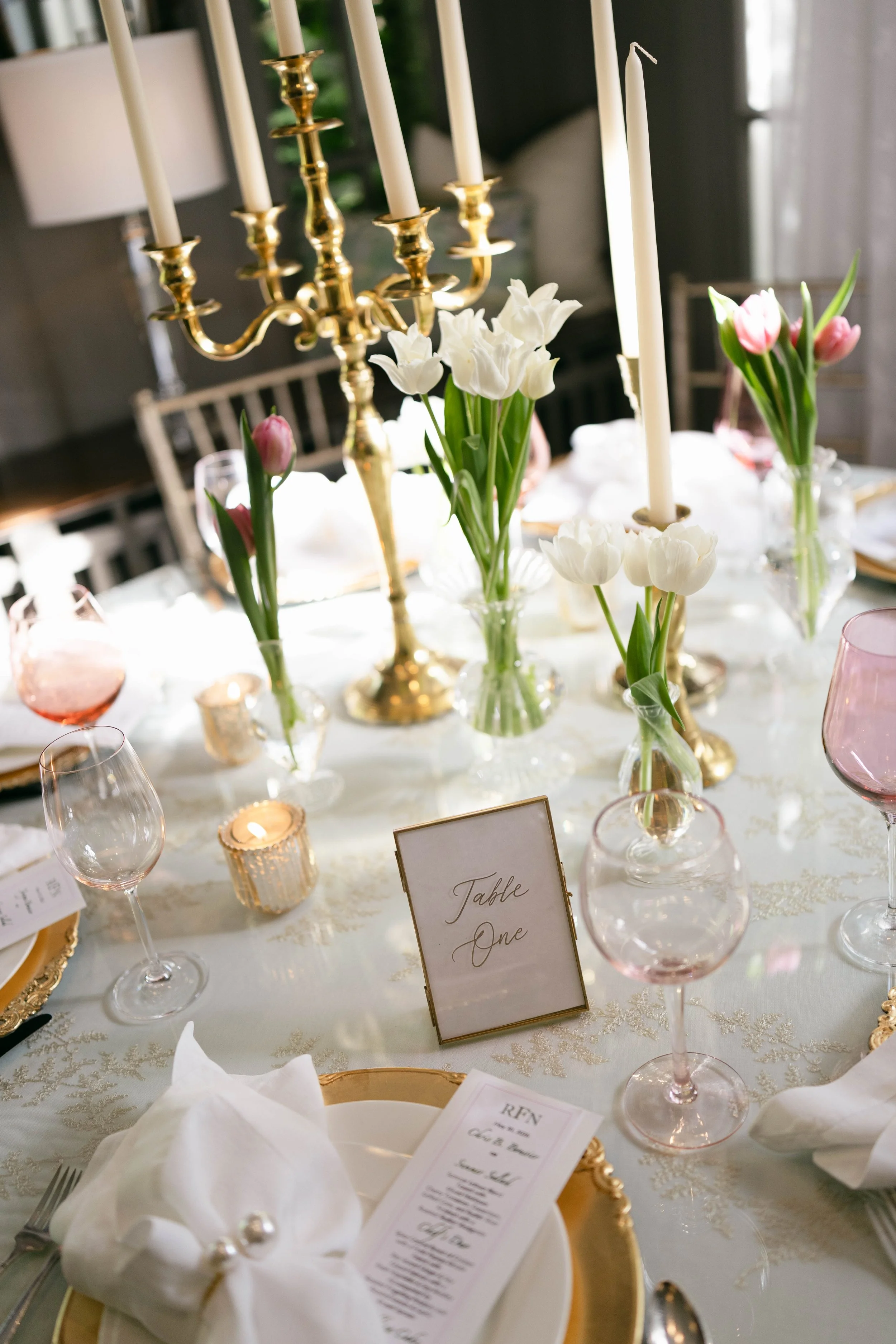 A formal dining table decorated with white and pink tulips in glass vases, a gold candelabrum with white candles, small candles, and a table number card labeled 'Table One'. The table is set with glassware, plates, and napkins.