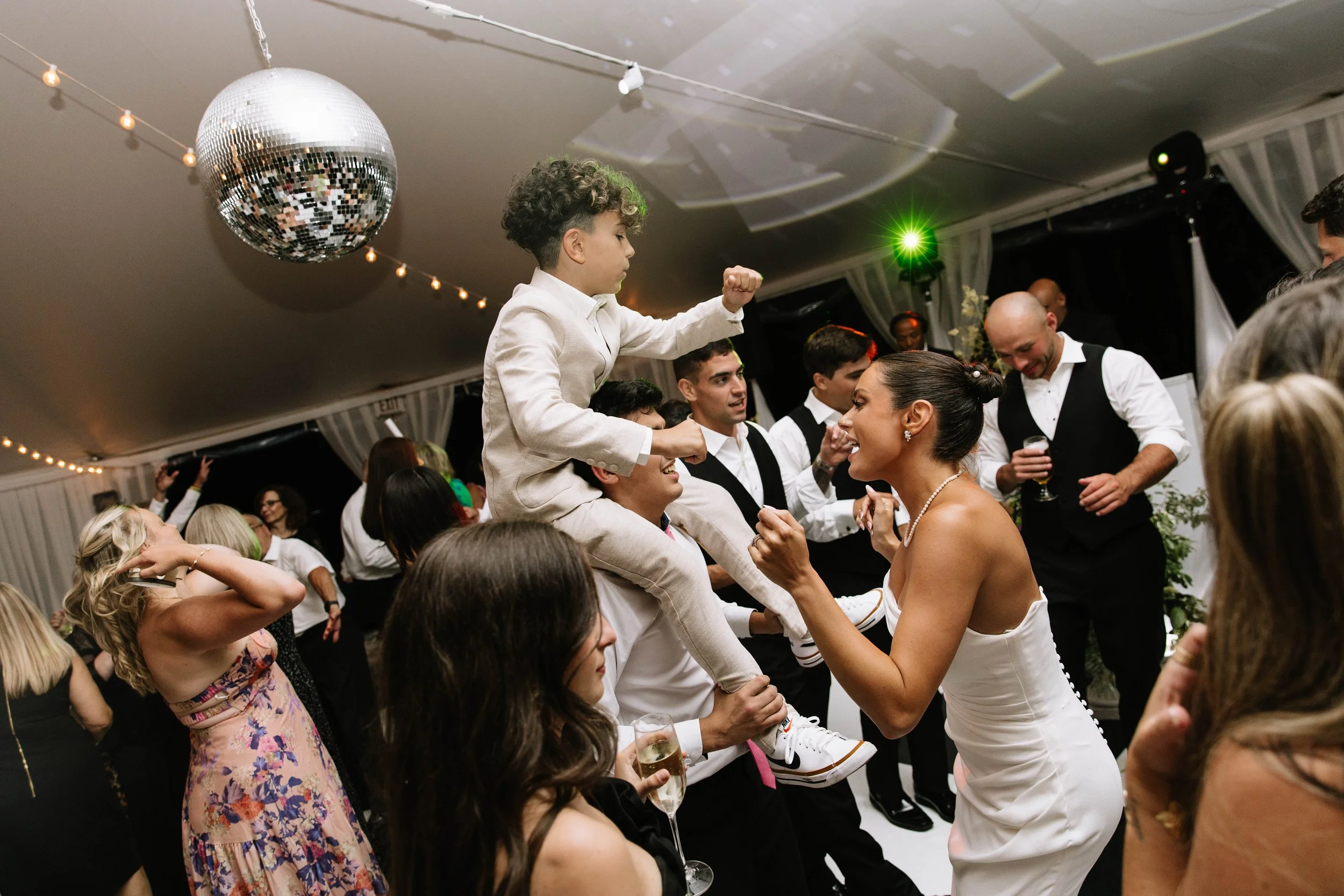 A lively celebration at a wedding reception with people dancing, smiling, and socializing under a decorated tent ceiling with string lights and a disco ball.