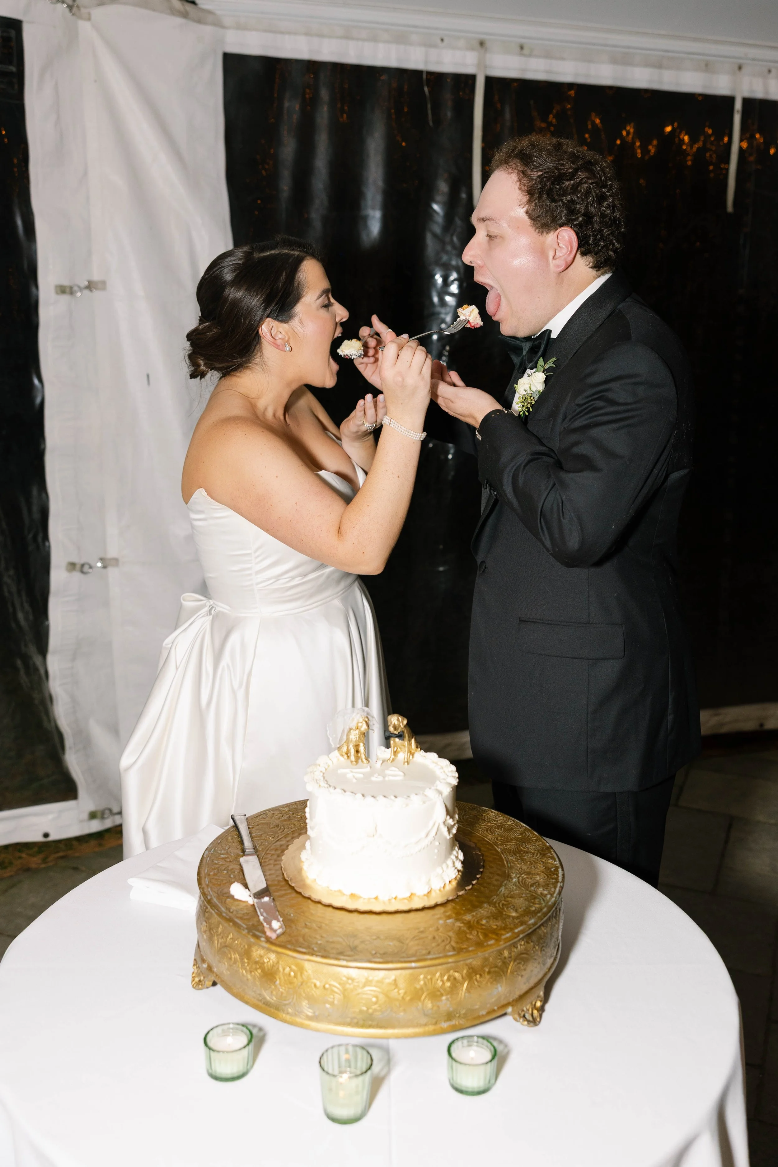 A bride and groom sharing cake at their wedding reception, with the bride in a white wedding dress and the groom in a black tuxedo, standing at a table with a white cake decorated with gold accents.