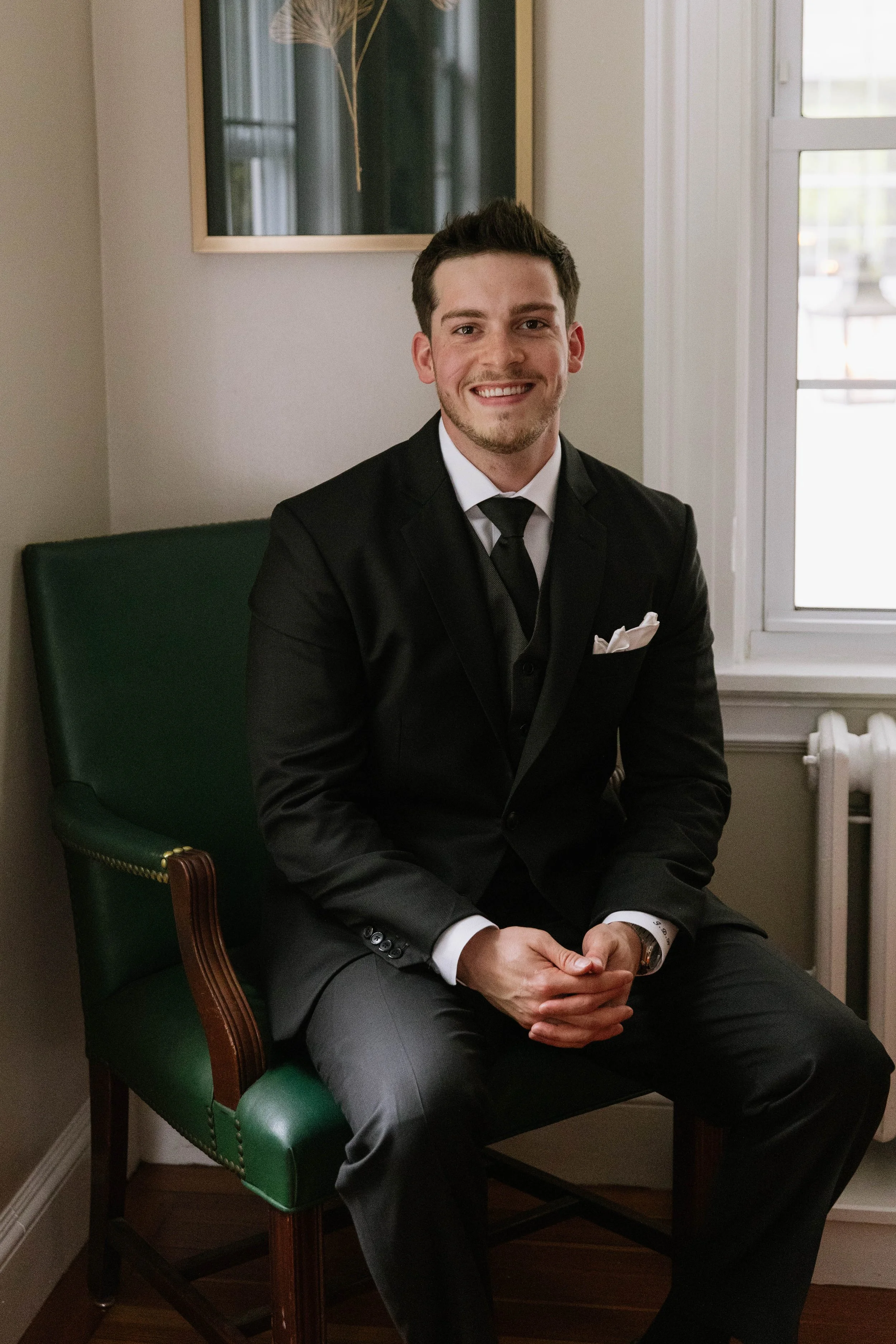 A smiling man in a black suit and tie sitting on a green chair in a room with a window and a piece of artwork on the wall.