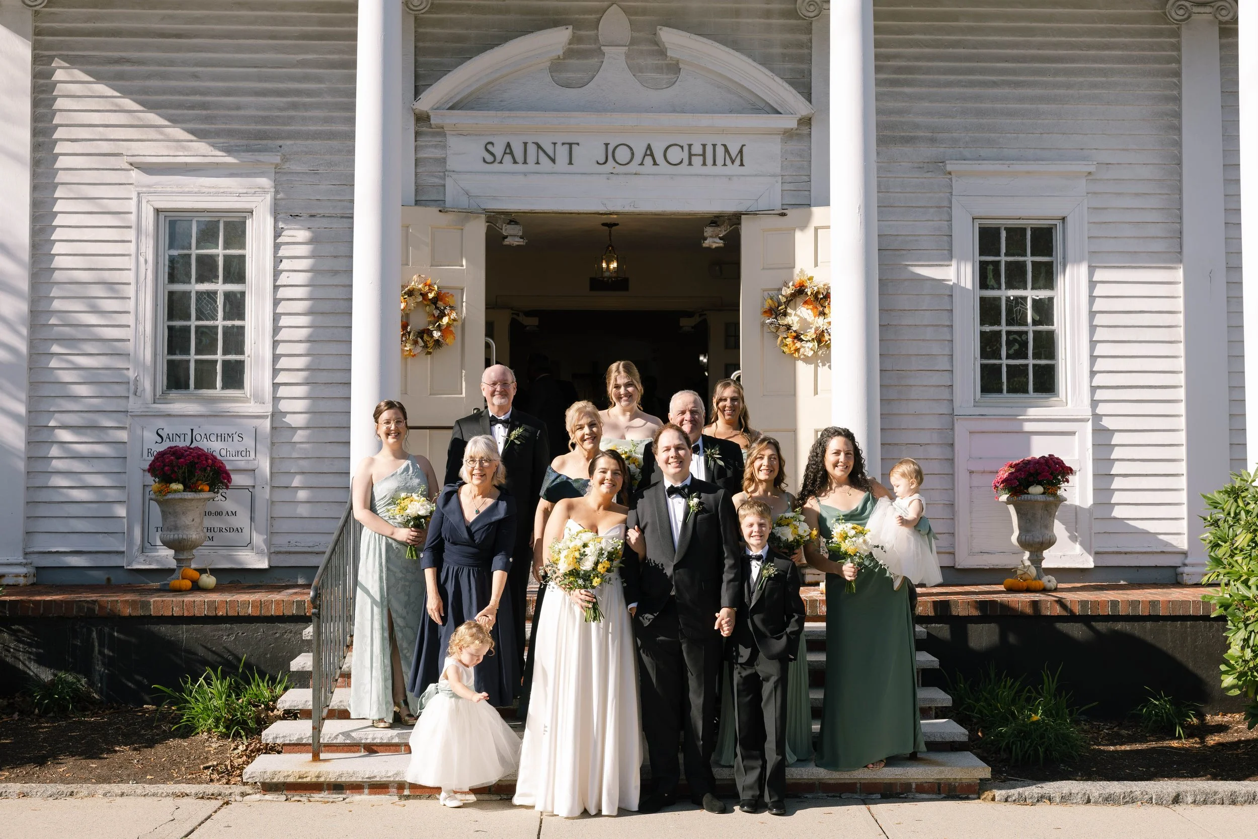 A wedding group photo in front of Saint Joachim's church showing a bride in a white dress, groom in a black tuxedo, bridesmaids and groomsmen, and family members, all smiling for the camera.