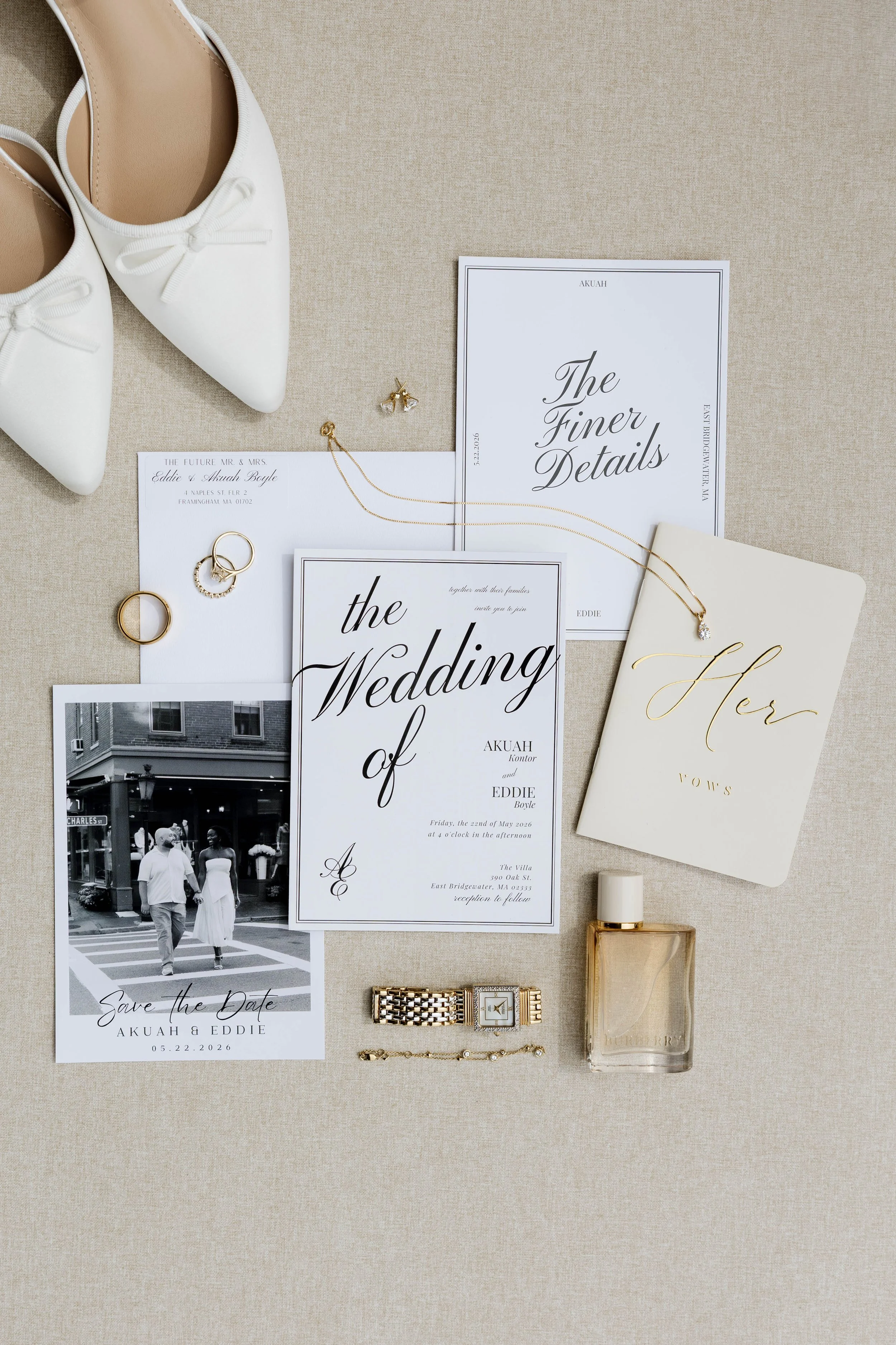 Wedding planning items including white shoes, jewelry, save the date card, wedding invitation, perfume, and a gold watch and bracelet, arranged on a beige fabric surface.