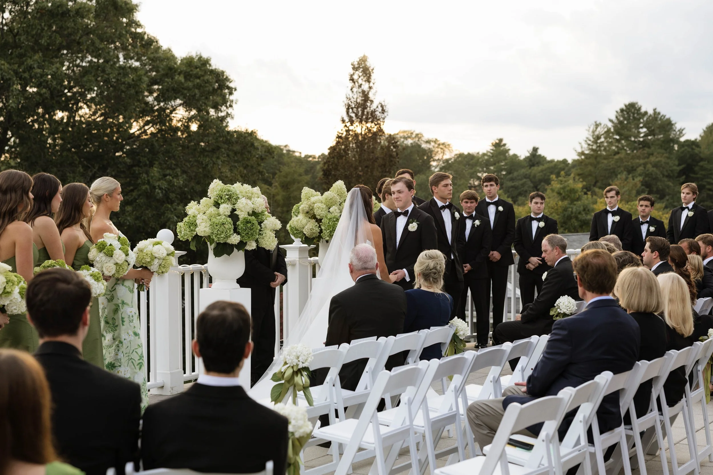 Outdoor wedding ceremony with bride and groom holding hands, surrounded by bridesmaids in green dresses and groomsmen in black tuxedos, with seated guests watching, in a lush garden setting.