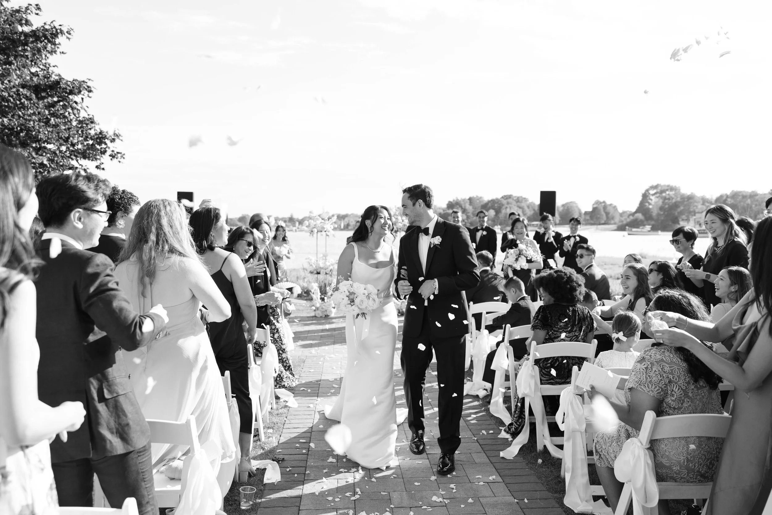 A black and white photo of a wedding ceremony outdoors with a bride and groom walking down the aisle, surrounded by guests seated and standing.