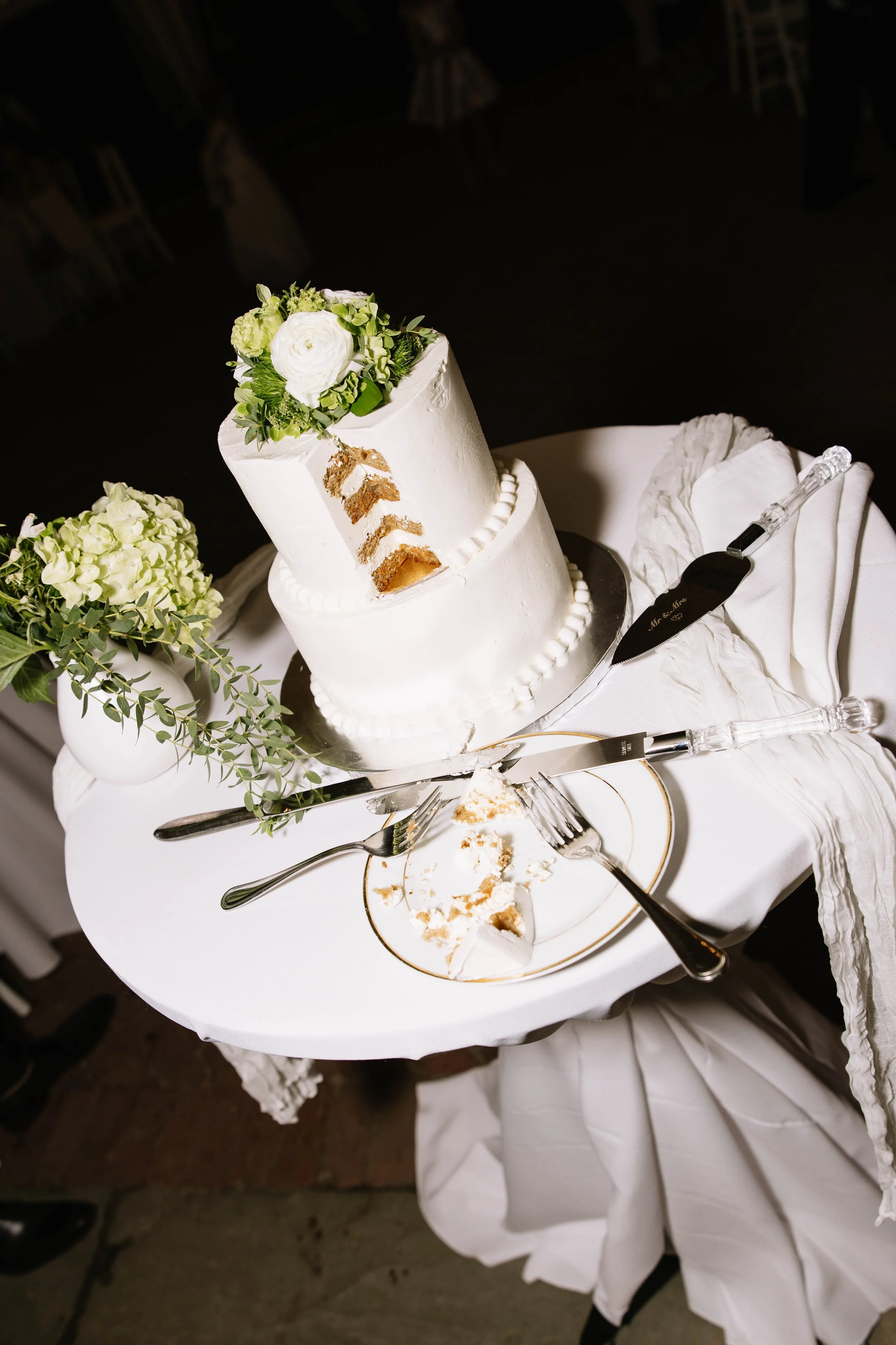 A wedding cake with white icing and floral decorations on top, placed on a white tablecloth with a cake knife and server, and a plate with cake crumbs and a slice missing.